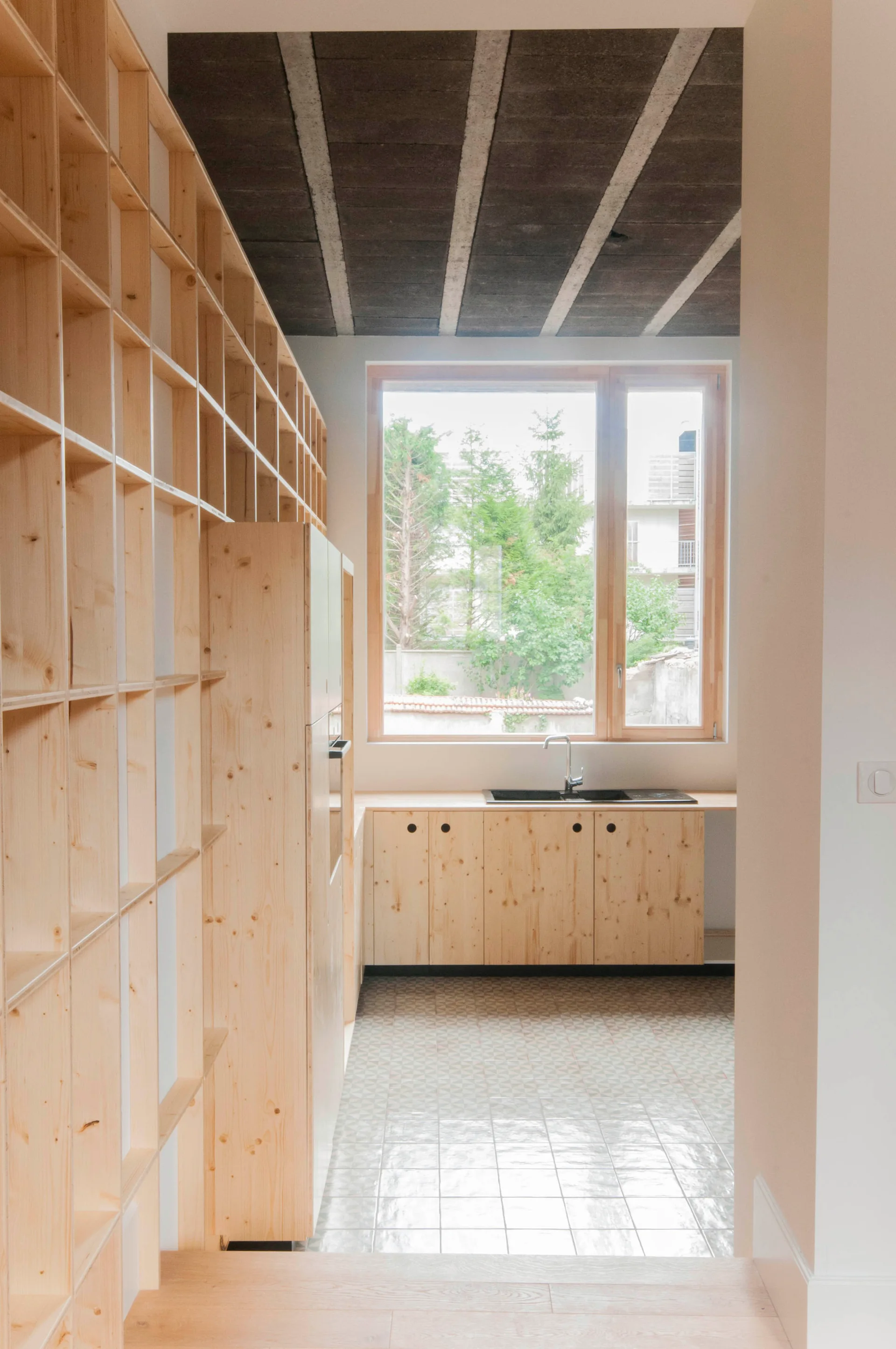 A minimalist kitchen features a large light-wood grid shelving unit, matching knotty pine cabinets under a window, and a tiled floor.