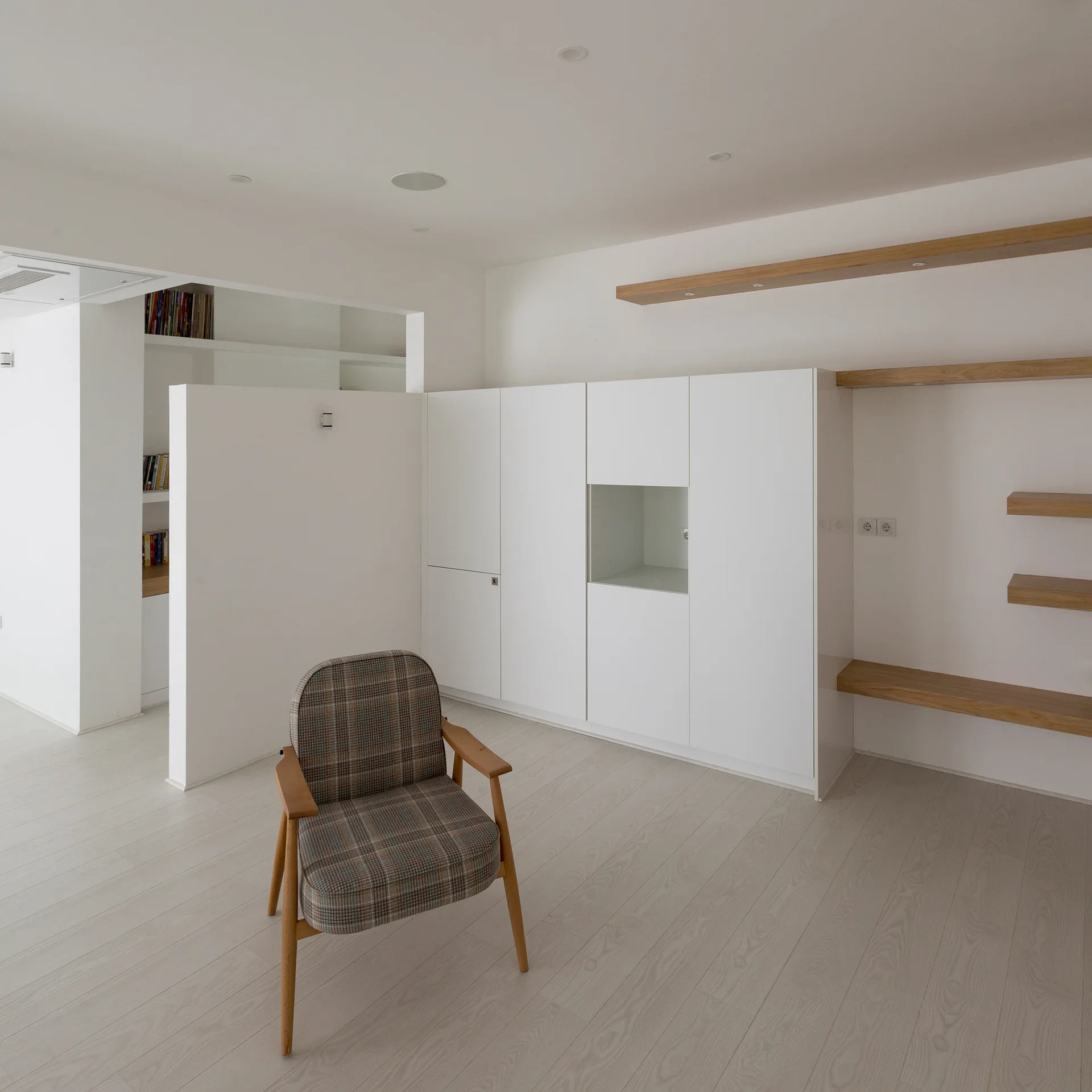 A plaid armchair sits on light wood flooring in a white minimalist room with built-in storage cabinets and thick oak wood shelves.