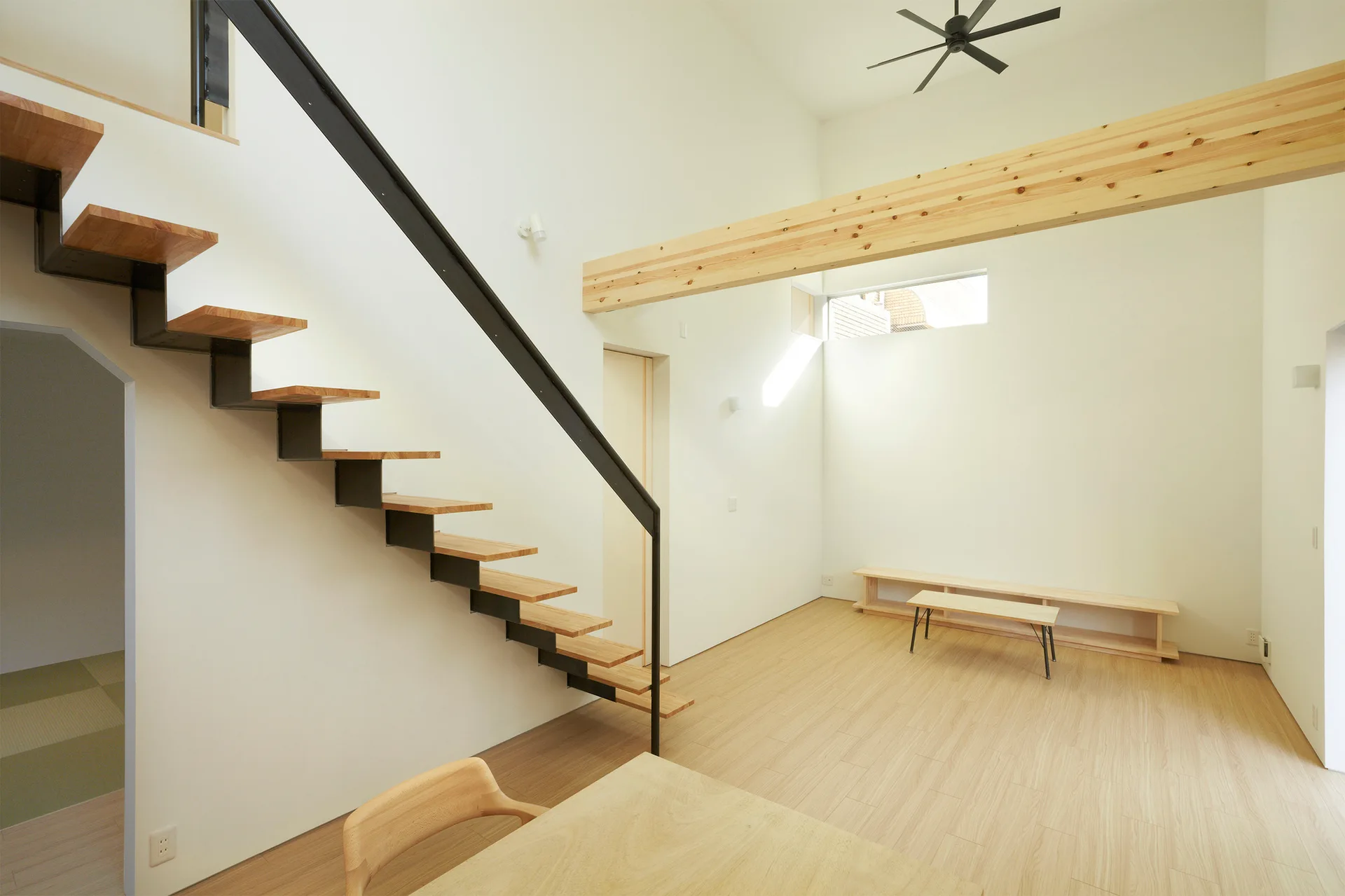 A minimalist living room features a floating wood staircase with a black metal frame, light wood floors, and an exposed timber ceiling beam.