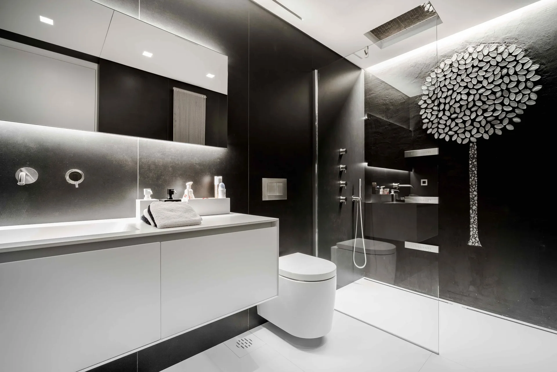 A contemporary bathroom features a white floating vanity, a wall-hung toilet, dark black tiled walls, and a glass-enclosed walk-in shower.