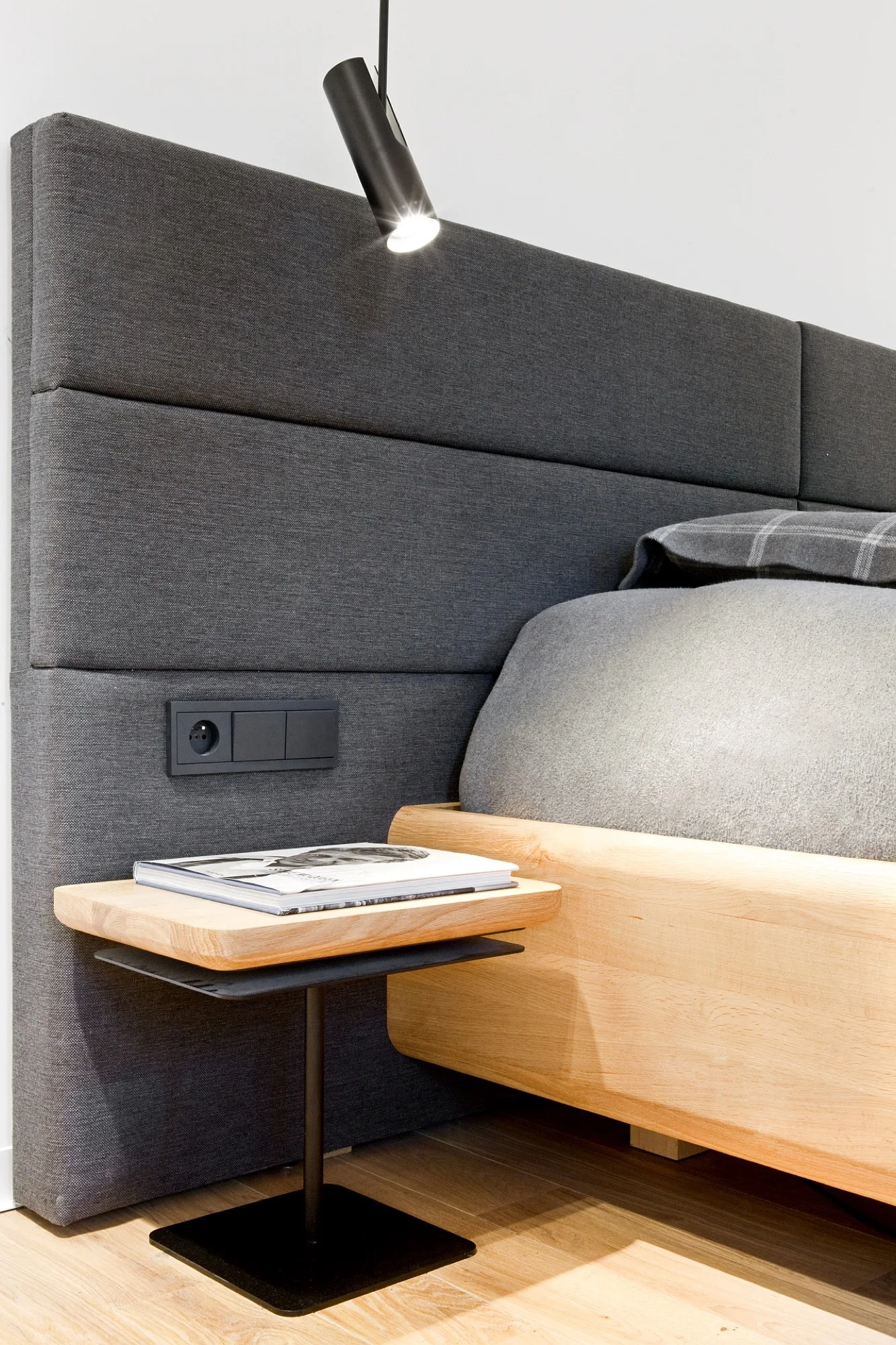 A close-up of a gray fabric headboard featuring a built-in black electrical outlet, a black spotlight, and a light wood bed frame.