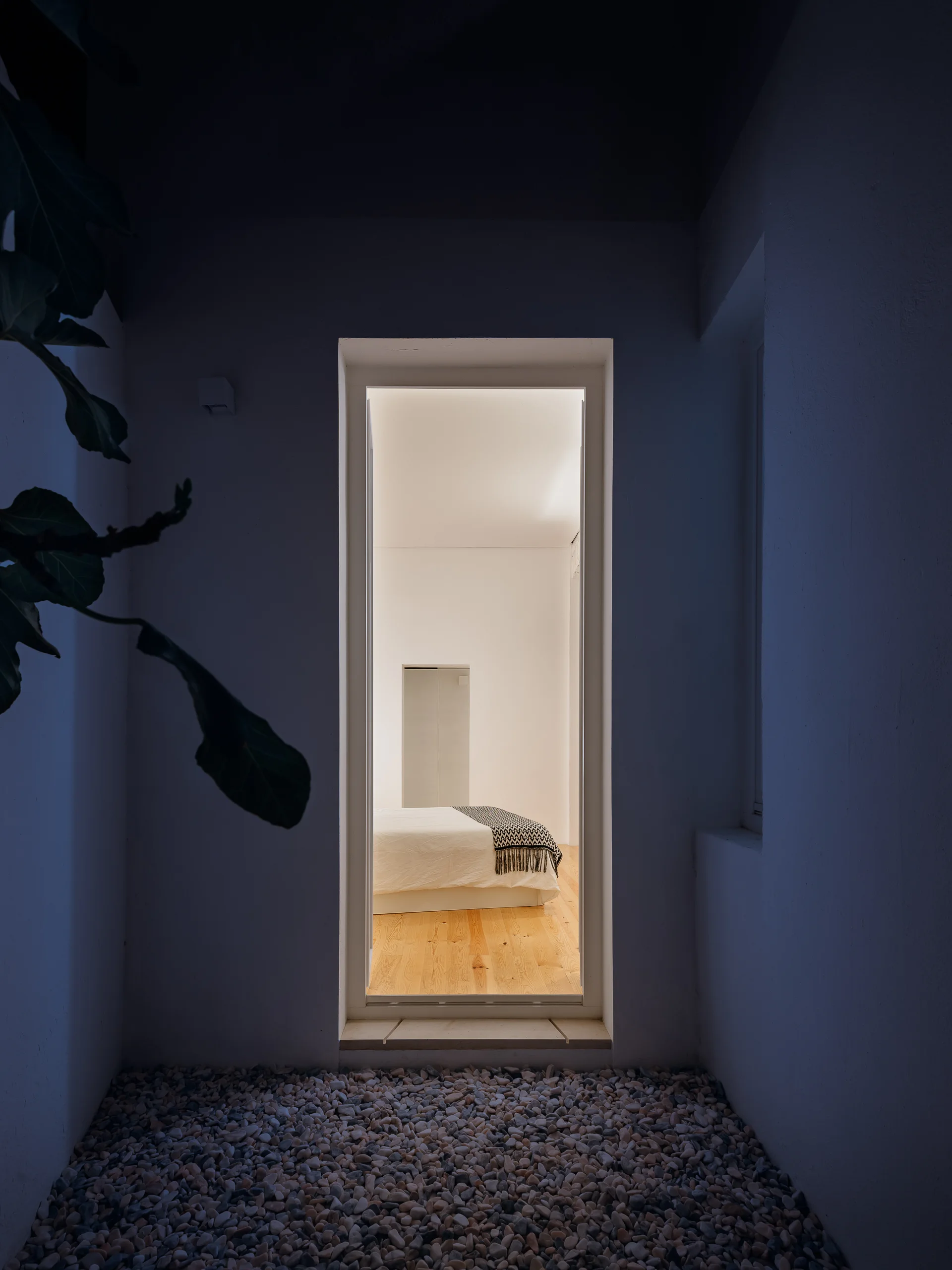 A view from a dark pebbled courtyard looking through a glass door into a brightly lit minimalist bedroom with wood floors.