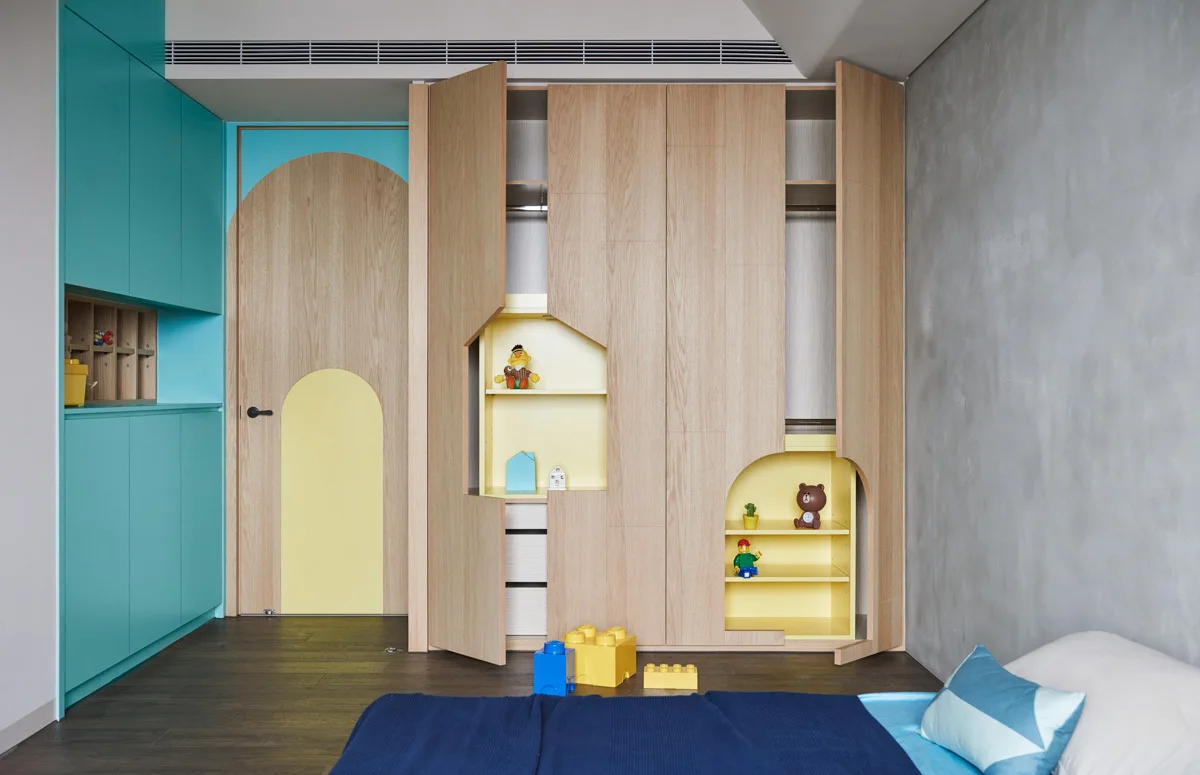 A children's bedroom features custom light wood wardrobes with arched cutouts, integrated yellow display shelves, teal cabinets, and a concrete-look wall.