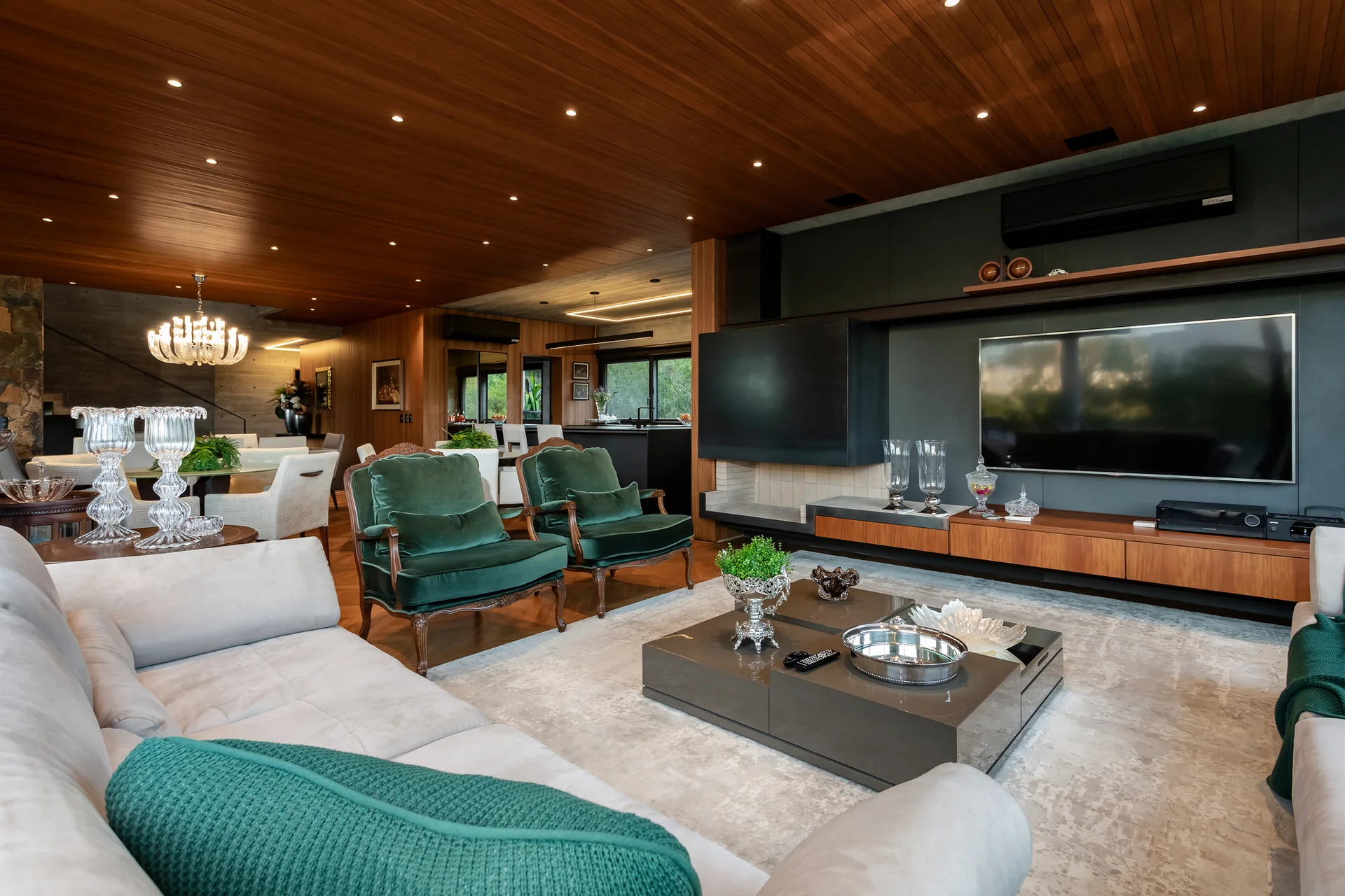 An open-concept living room features a warm wood-planked ceiling, a light grey sectional sofa, and two emerald green velvet armchairs.
