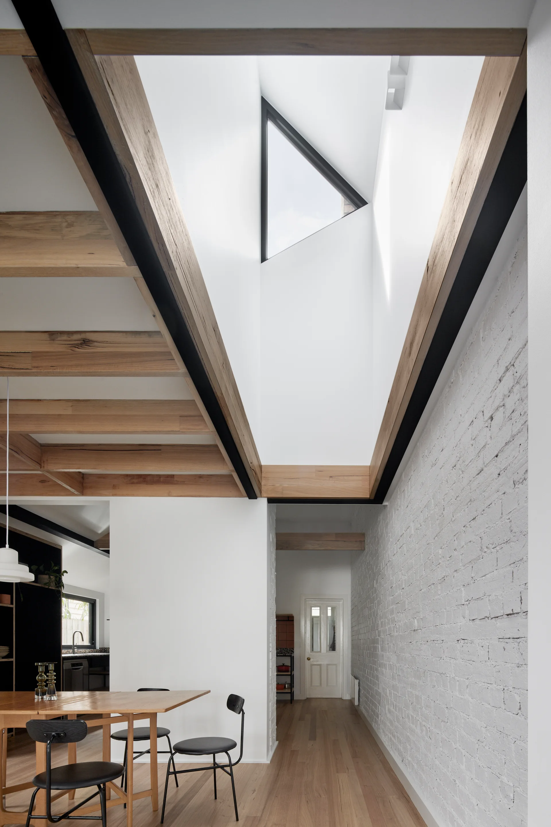A modern dining room features a wooden table with black chairs, a white painted brick wall, and exposed wooden ceiling beams below a triangular skylight.