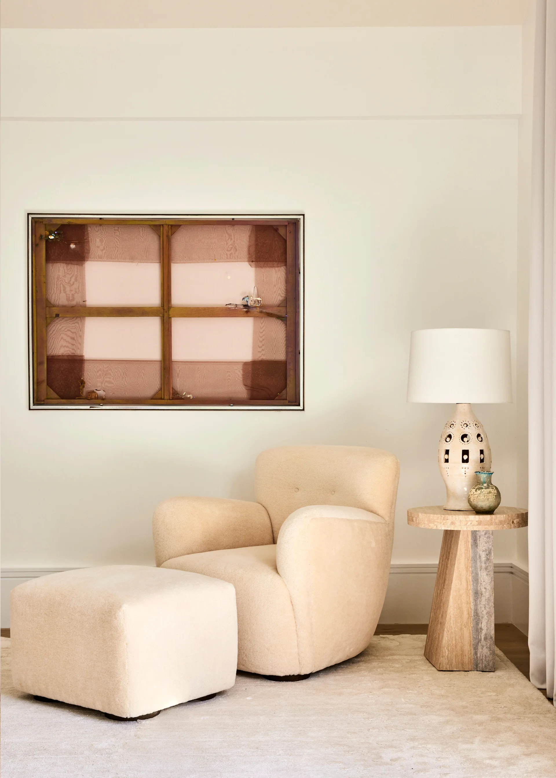 A cream upholstered armchair and matching ottoman sit next to a travertine pedestal side table with a ceramic lamp.