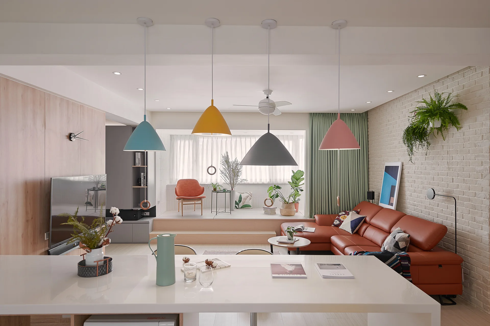 A modern open living room with colorful pendant lights, a brown leather sectional, a white brick wall, and an elevated wooden platform.