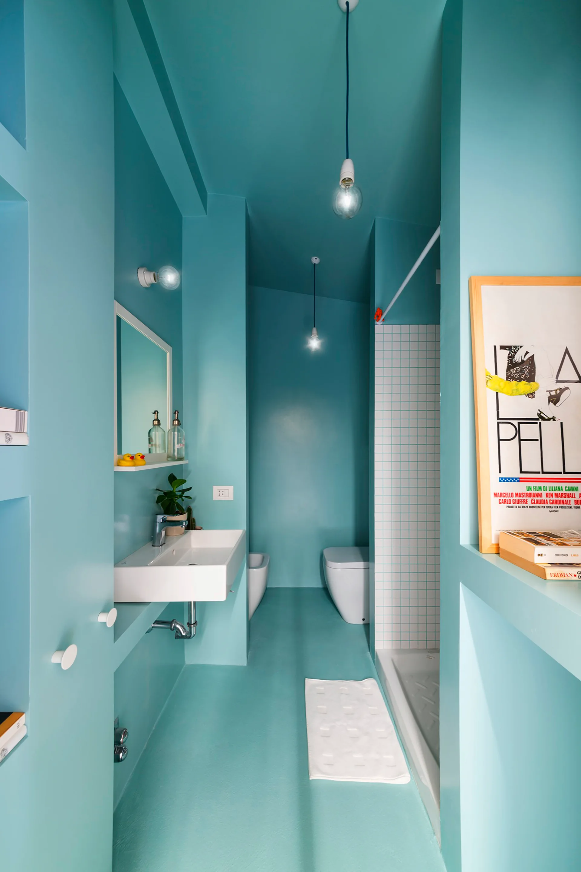 A narrow bathroom features turquoise walls, ceiling, and floor with a white wall-mounted sink, a toilet, recessed shelving, and a tiled shower stall.