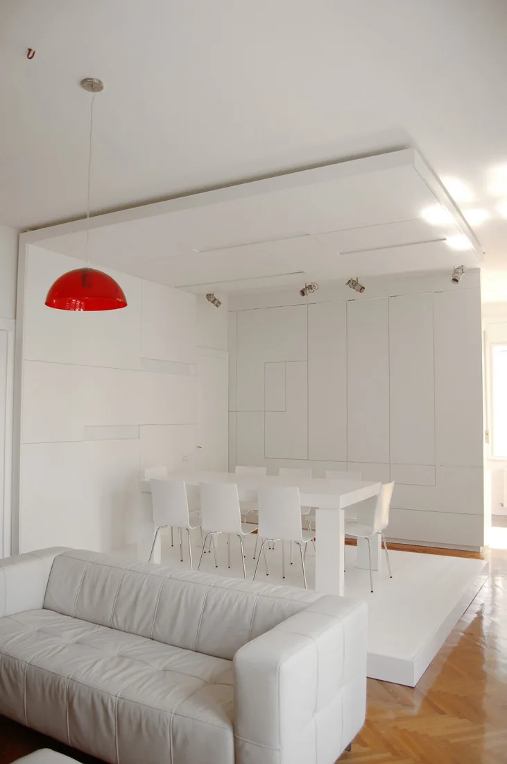 A minimalist white dining room features a table and chairs on a raised white platform next to a modern tufted sofa and a red pendant light.
