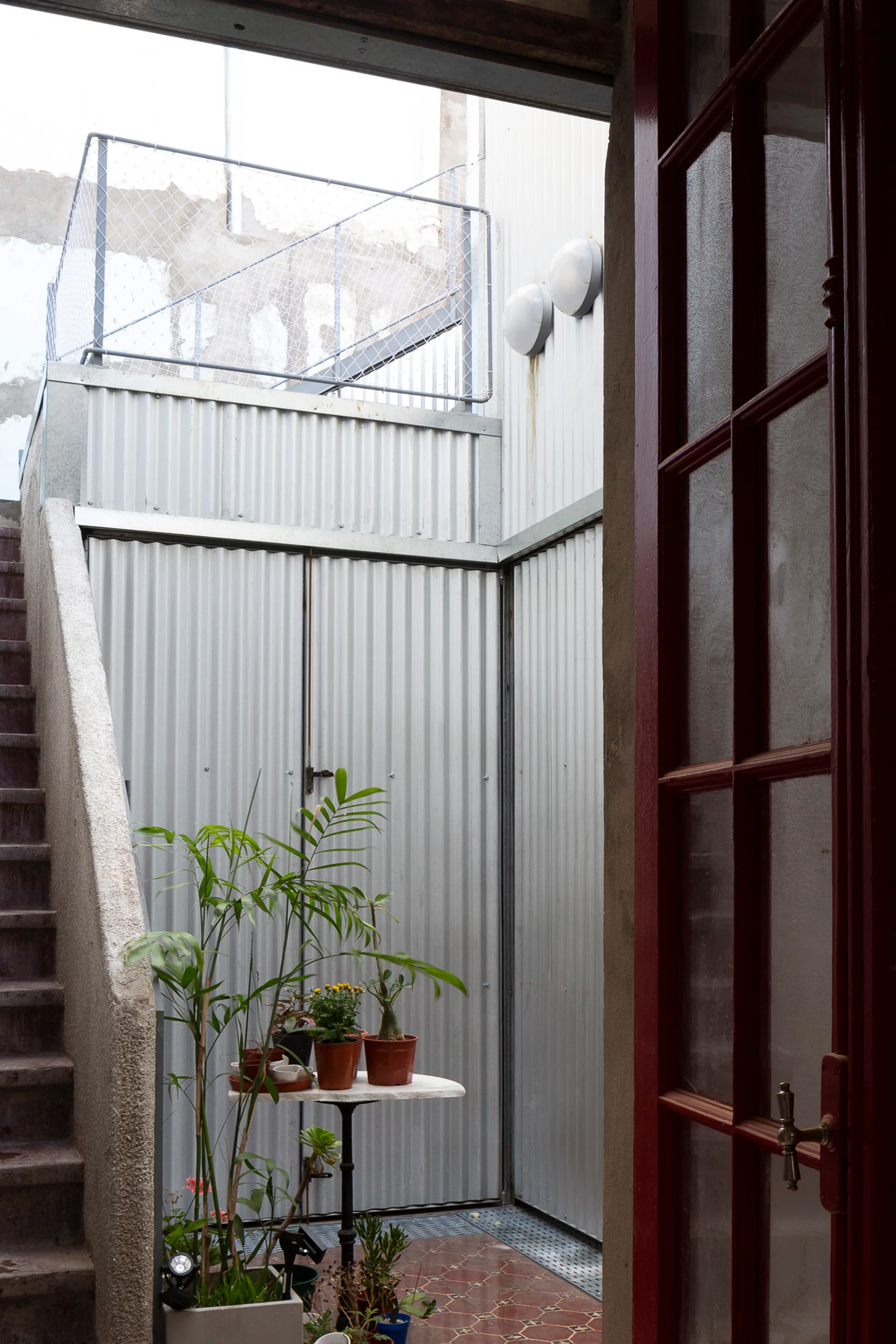 A view framed by a red door shows an industrial courtyard with corrugated metal walls, concrete stairs, and a small table holding potted plants.