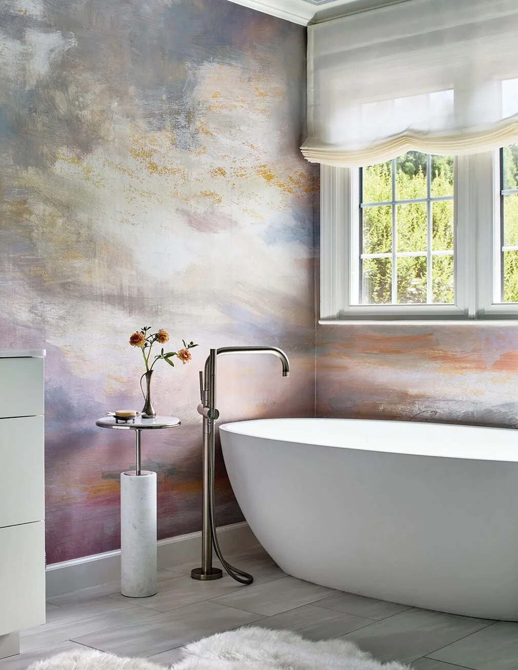 A white freestanding bathtub sits next to a colorful, painterly mural wall, a marble side table, and a window with a white roman shade.