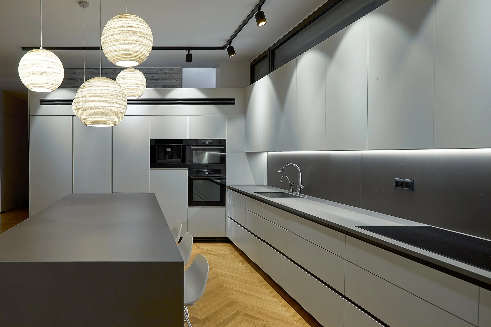 A minimalist kitchen features matte white cabinets, a large grey island, herringbone pattern wood flooring, and sphere pendant lights.