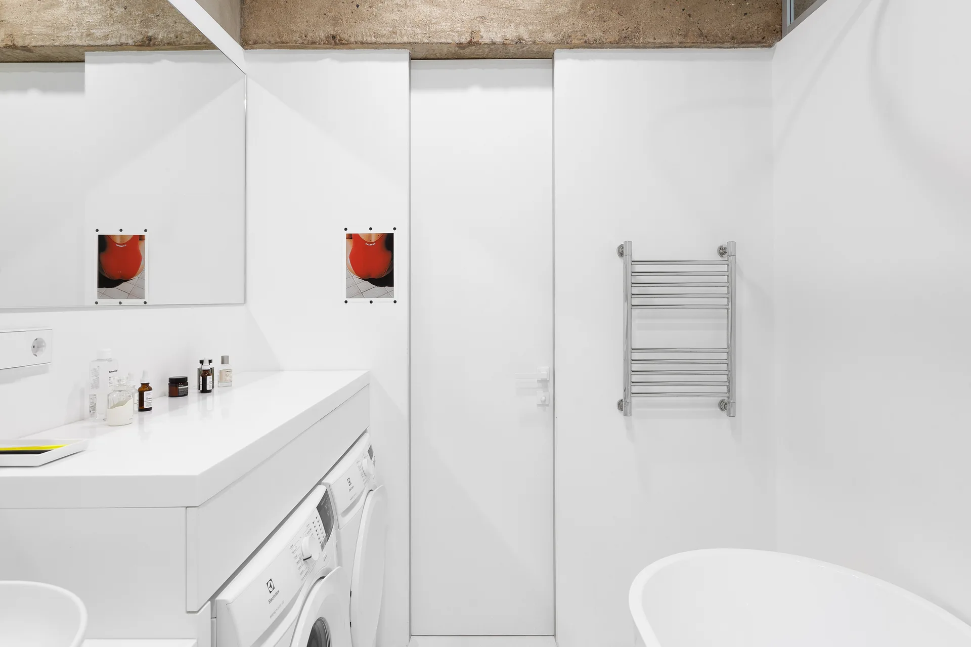 A minimalist white bathroom features laundry appliances built under a long vanity, an exposed concrete ceiling beam, and a chrome towel warmer.