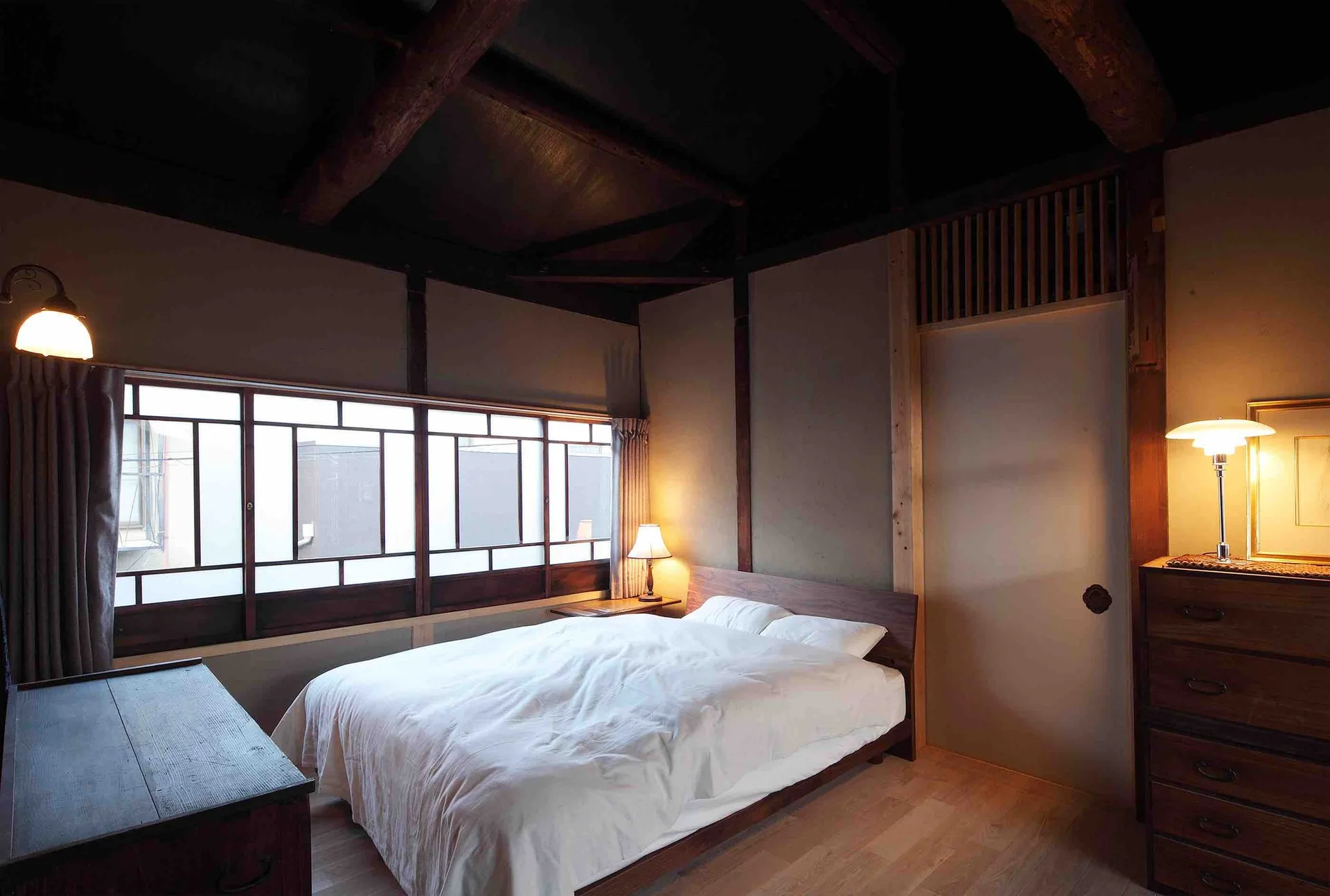A minimalist bedroom featuring a low wooden bed with white bedding, dark exposed ceiling beams, warm table lamps, and a horizontal lattice window.