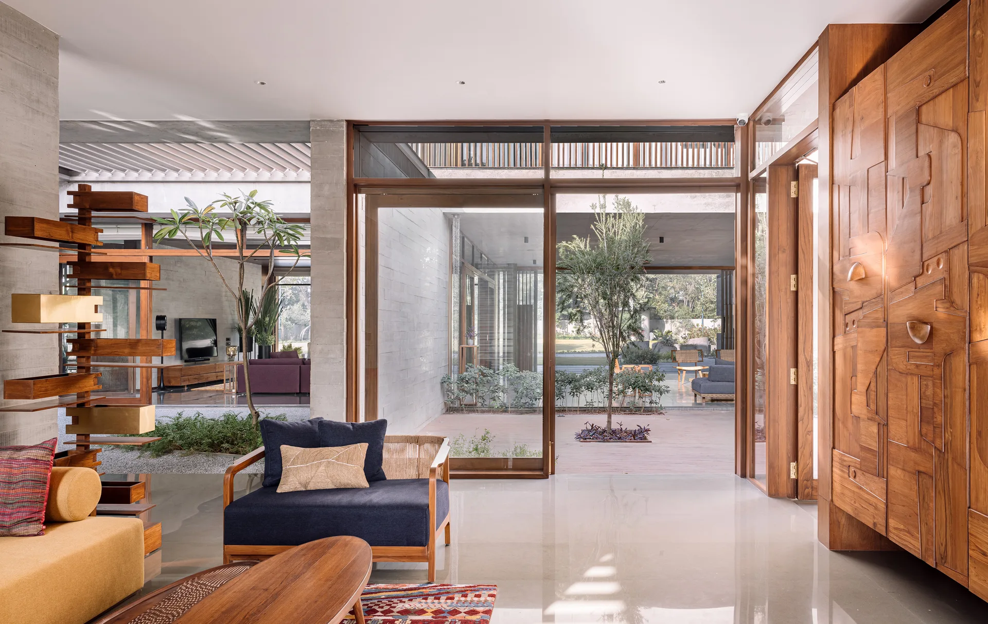 A contemporary living room featuring a carved wooden wall cabinet, a blue armchair, and large glass doors looking out onto a central courtyard.