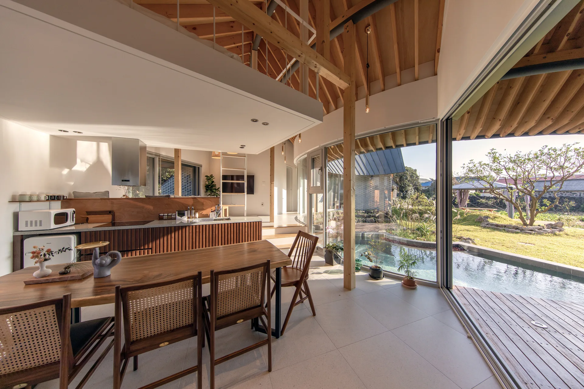 An open dining space features a wooden table with cane back chairs, a slatted wood kitchen island, and sliding glass doors opening to an outdoor pool.