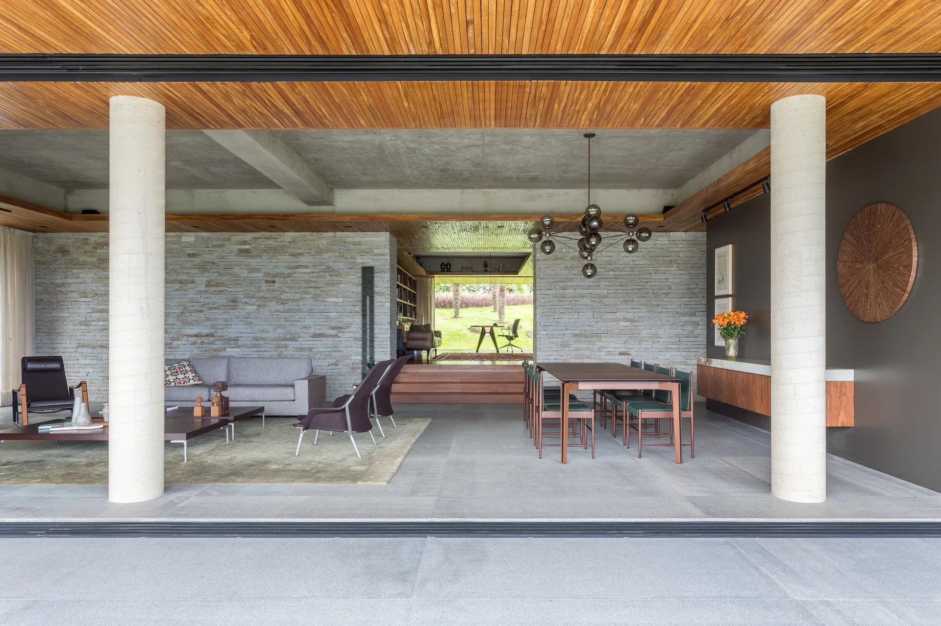 An open-plan living and dining space with concrete pillars, a slatted timber ceiling, a textured stone wall, and a modern dining table set.