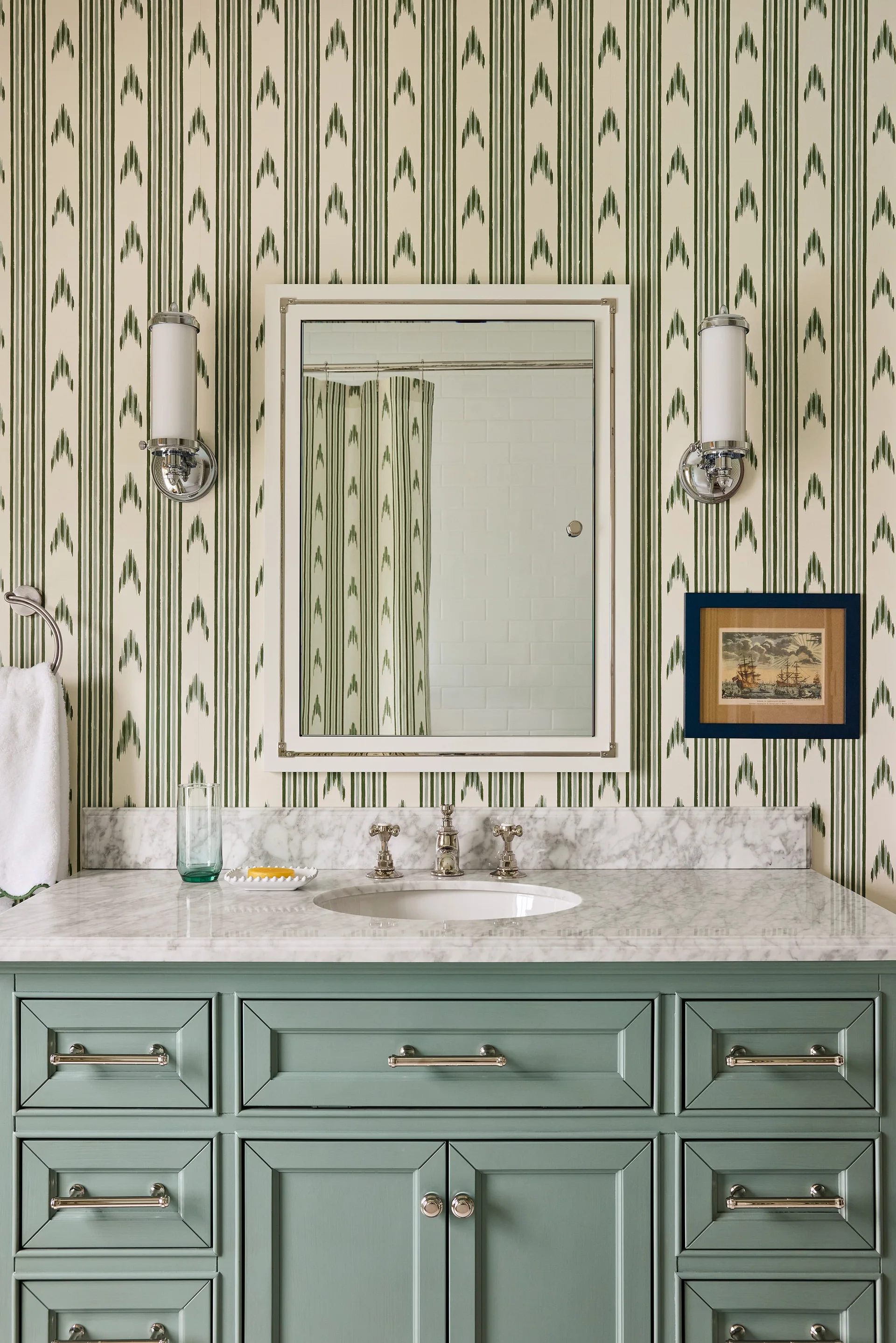 A sage green vanity with a marble countertop is set against green and cream striped wallpaper below a rectangular mirror and two silver sconces.