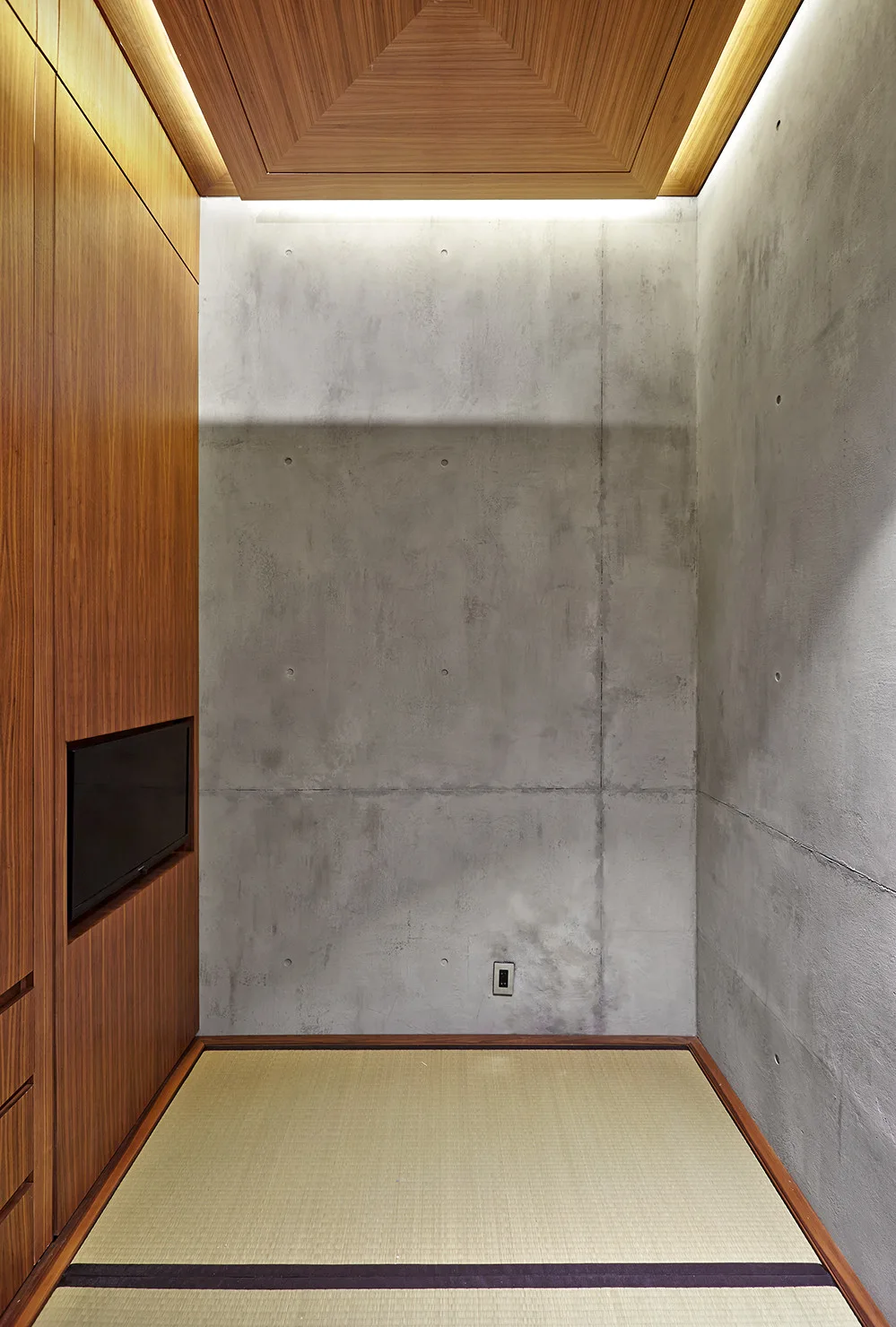 A minimalist room featuring traditional tatami mat flooring, smooth exposed concrete walls, and built-in wood cabinetry illuminated by soft cove lighting.