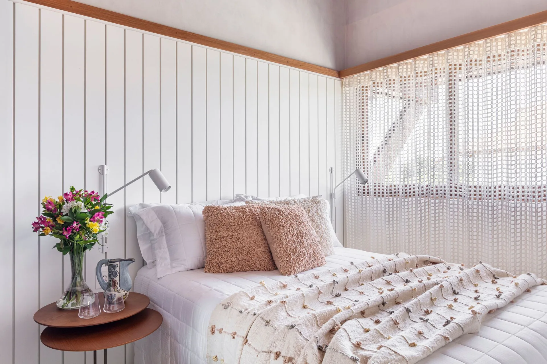 A bedroom featuring white vertical wood paneling, a bed with textured pink pillows, and a tiered wooden nightstand next to a grid-patterned curtain.