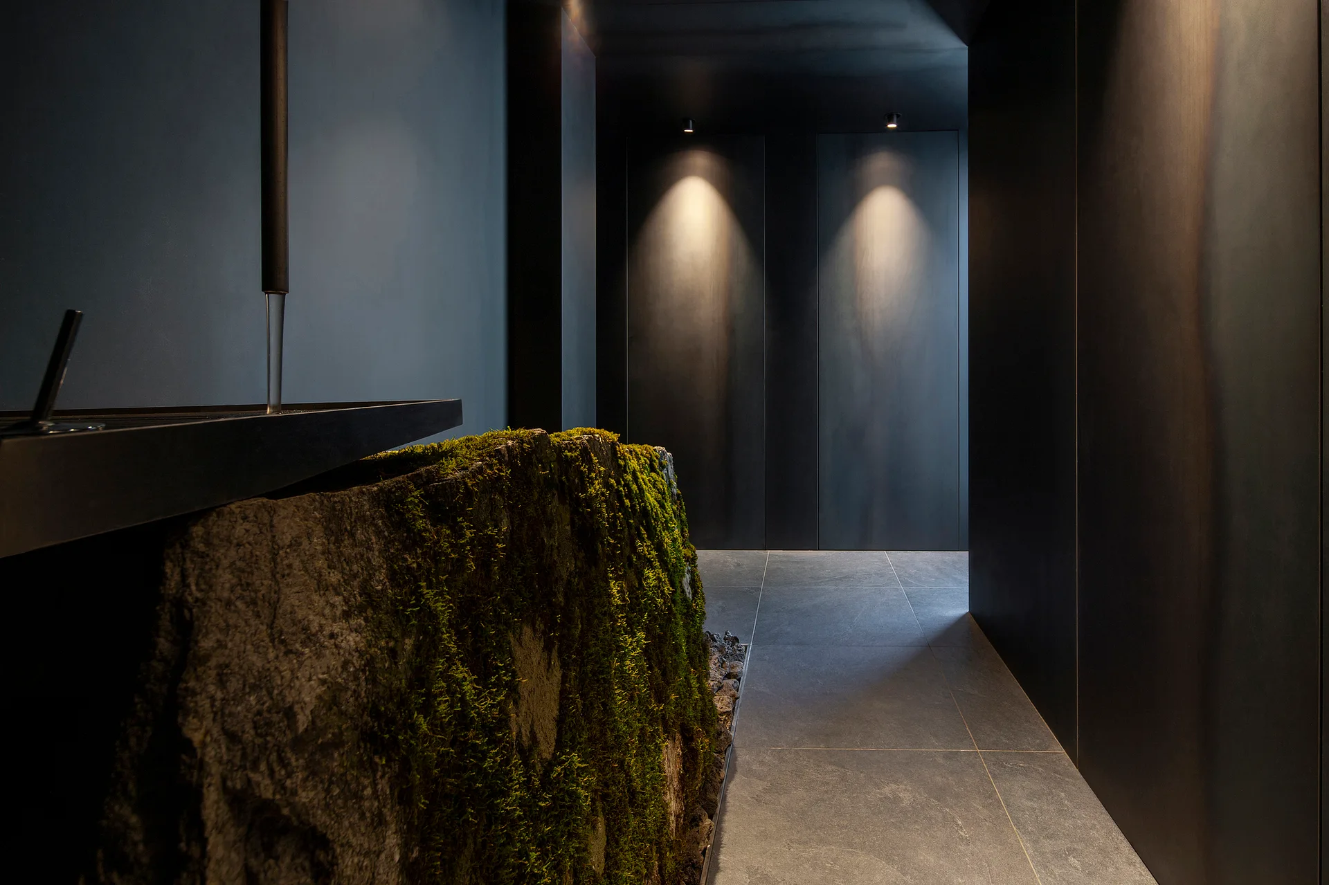 A dark minimalist hallway features matte black paneled walls, stone tile floors, and a large natural moss-covered stone sculpture lit by spotlights.