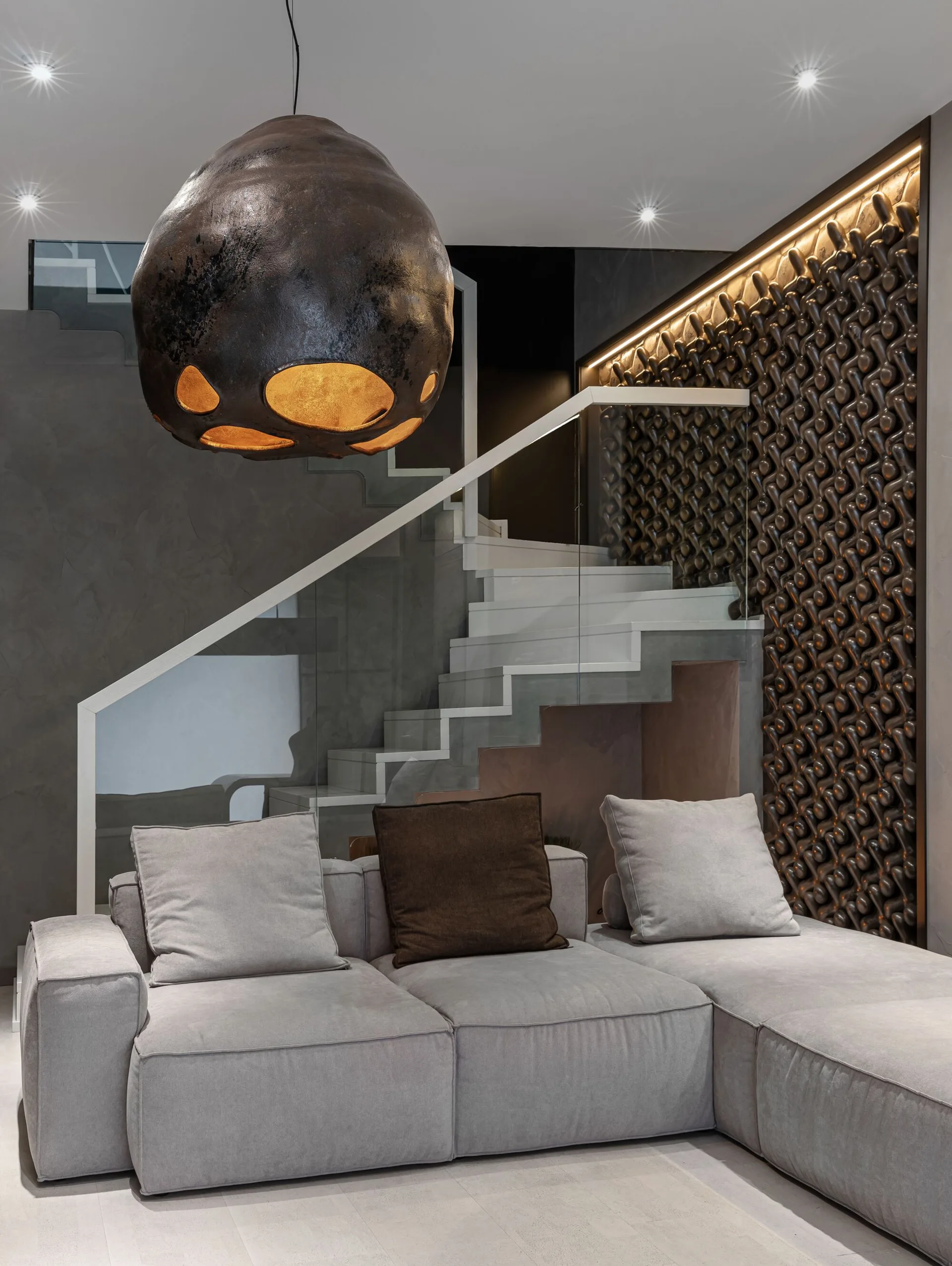 A modern grey sectional sofa sits near a staircase with a glass railing and a textured bronze accent wall illuminated by integrated overhead lighting.