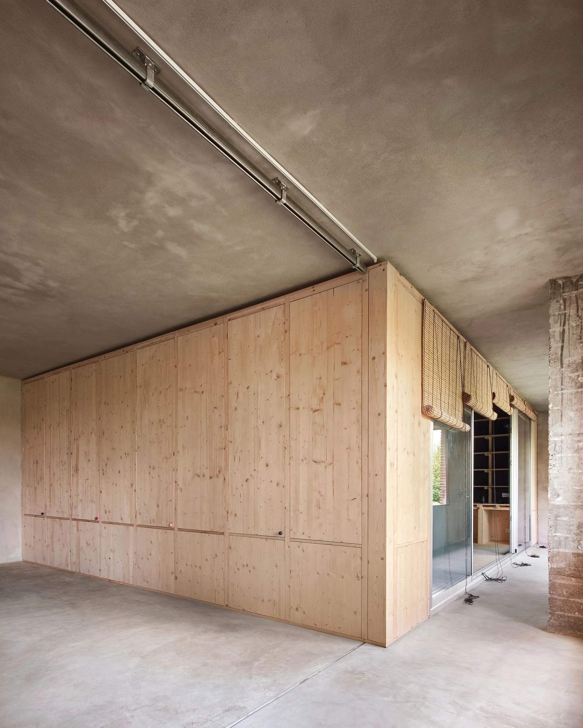 A minimalist interior featuring raw concrete floors and ceilings paired with a massive light wood built-in storage cabinet and sliding glass doors.