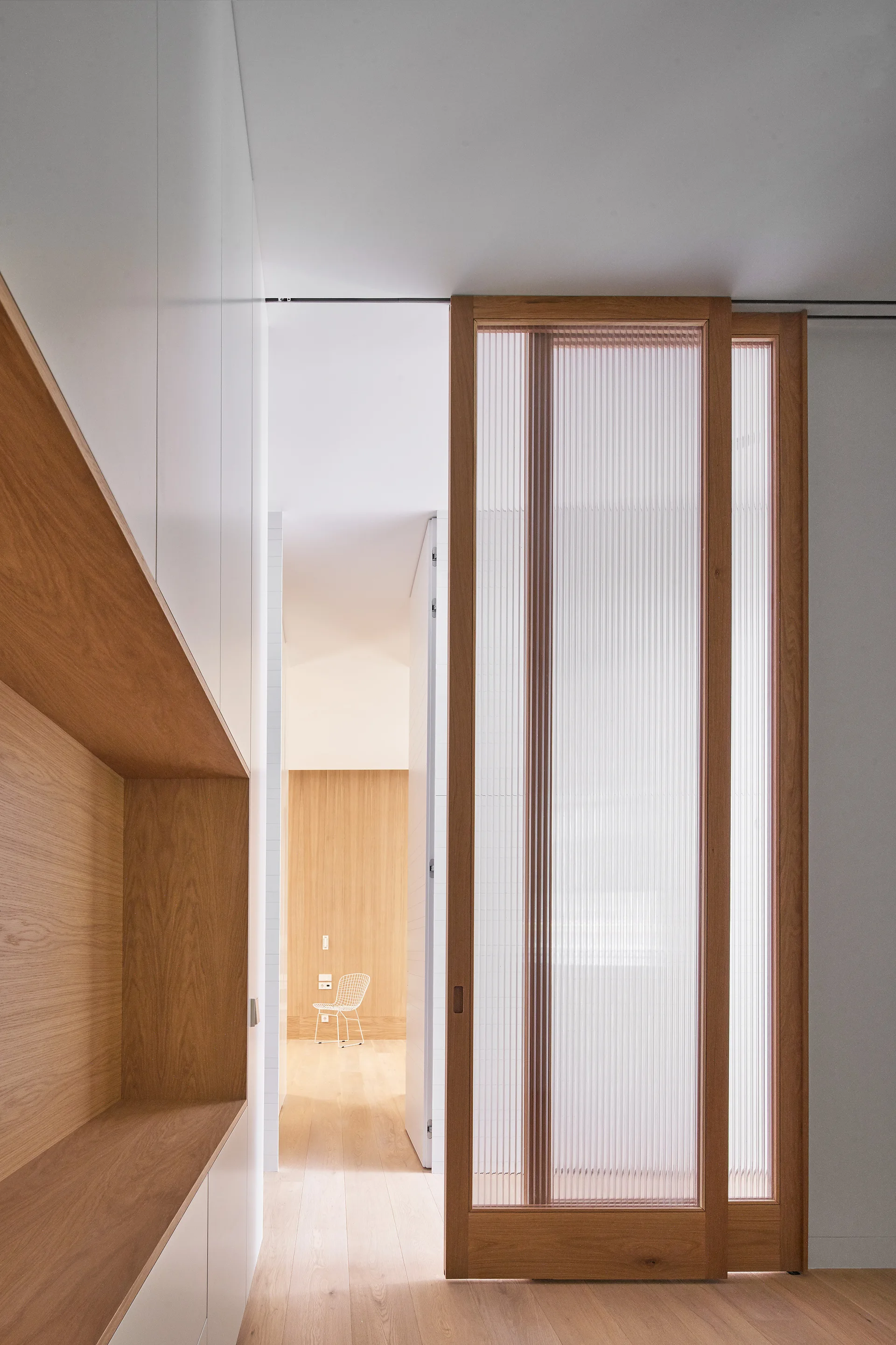 A minimalist hallway features a wooden-framed sliding door with fluted glass, white built-in storage with a wood niche, and light wood flooring.