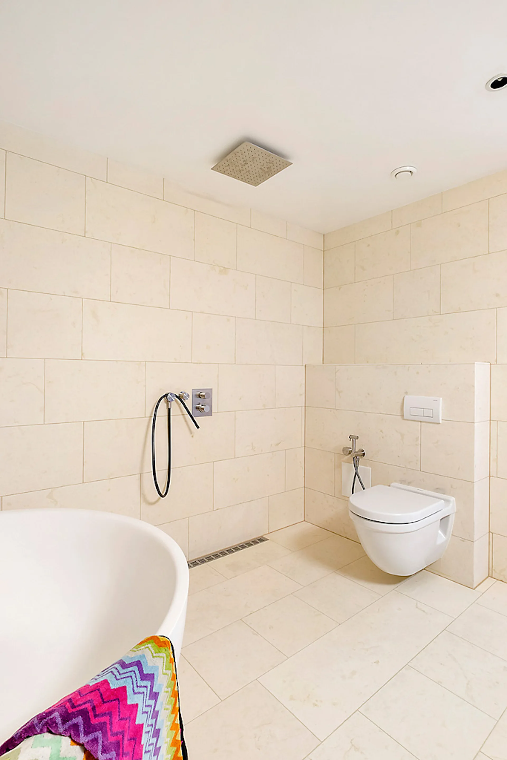 Modern bathroom with large beige stone tiles, a wall-mounted toilet, a linear floor drain, and a white soaking tub in the foreground.