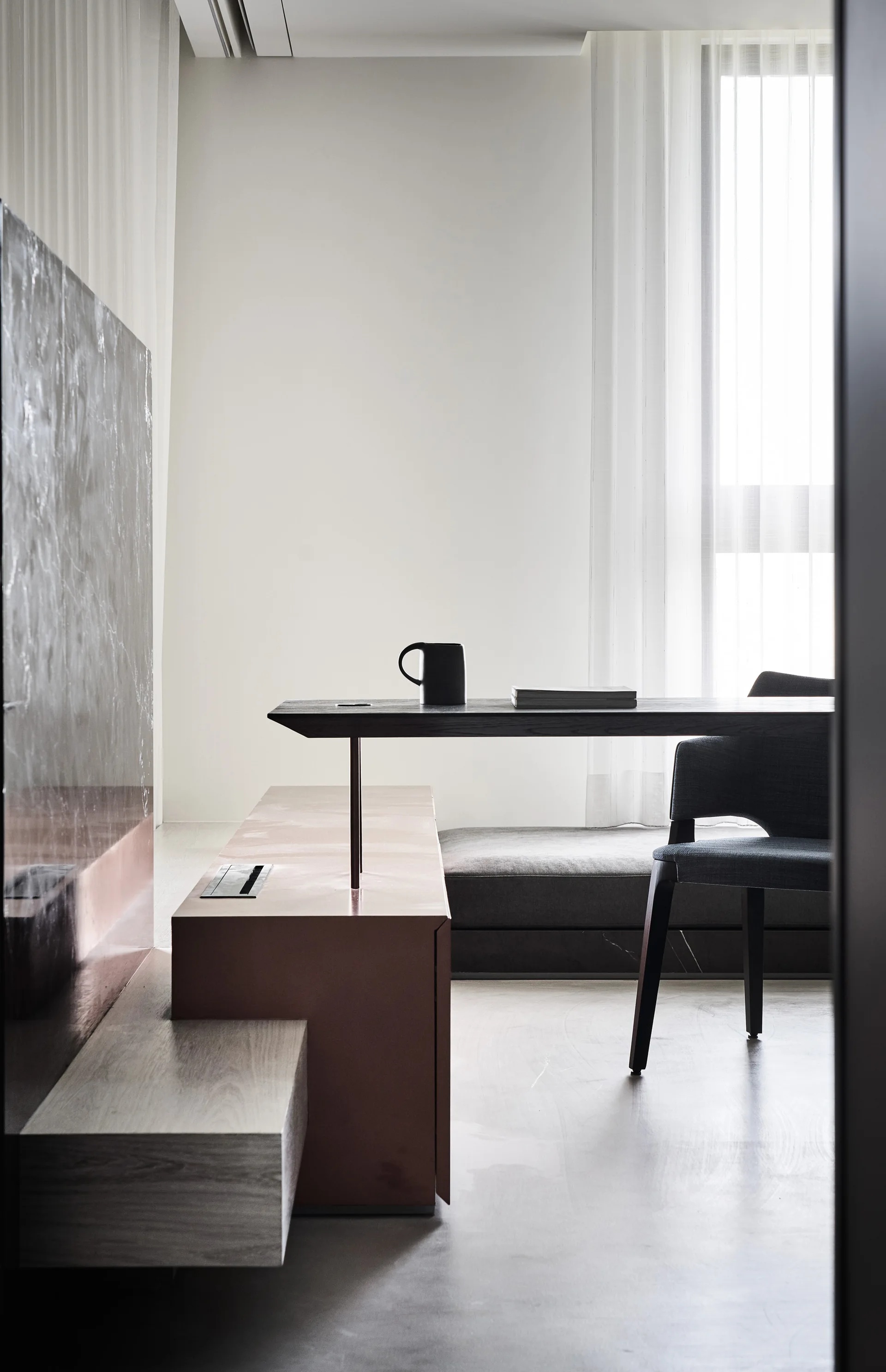 A minimalist home office features a slim dark desk with a black mug, a gray upholstered chair, and a low multi-level copper-toned bench by a window.