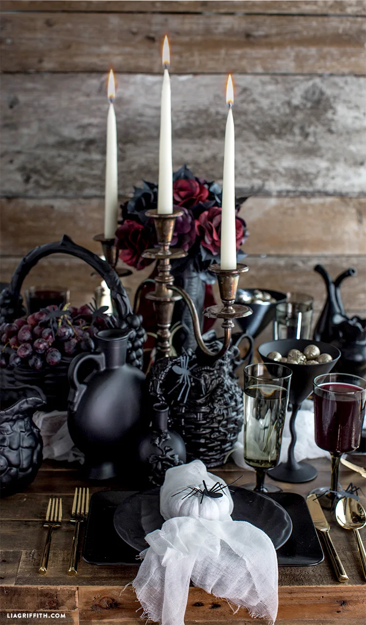 A dark table setting features a metal candelabra with lit candles, matte black ceramic plates, and gold flatware against a rustic wood backdrop.