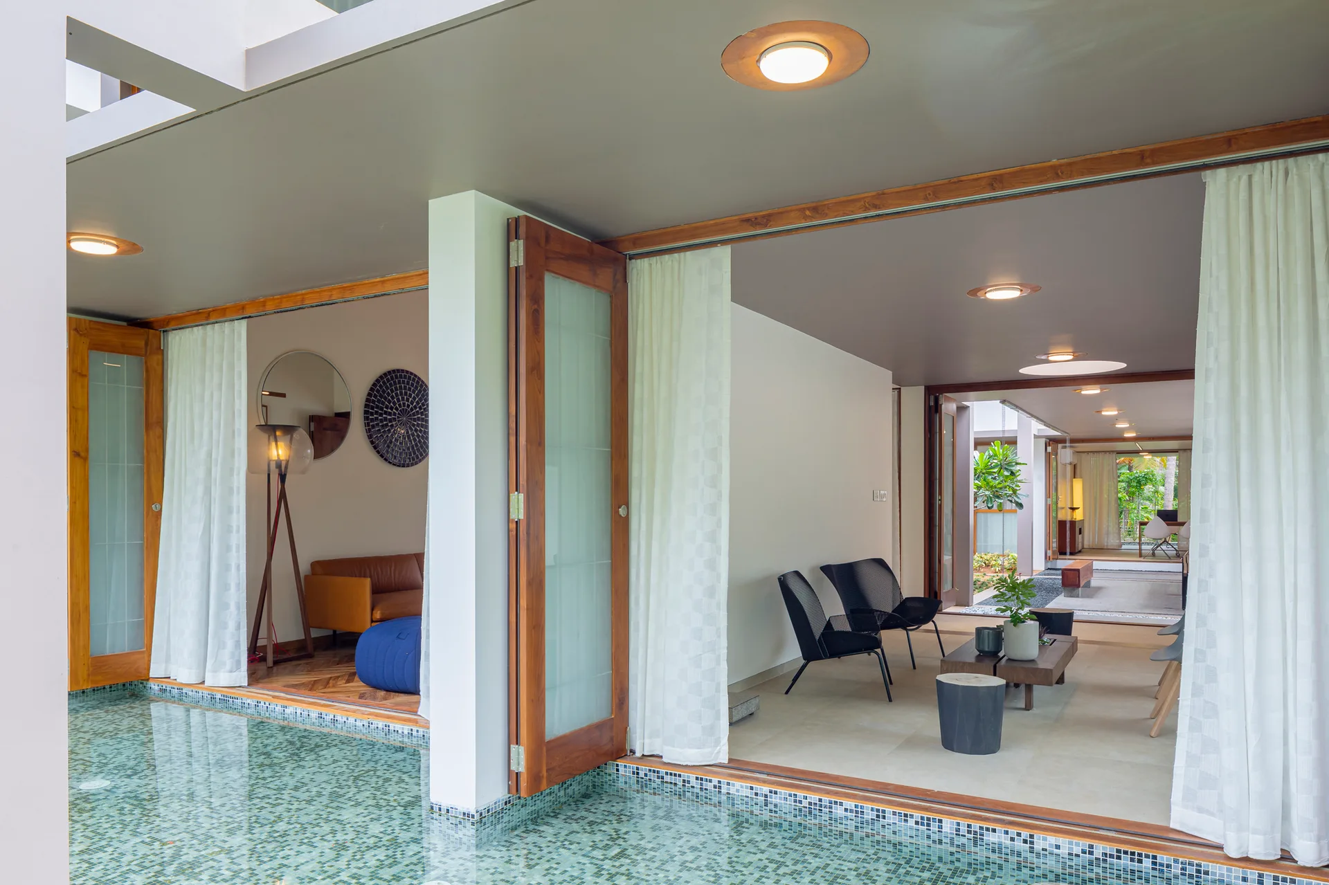 An open indoor-outdoor patio with black armchairs and wood folding doors adjacent to a blue tiled swimming pool.