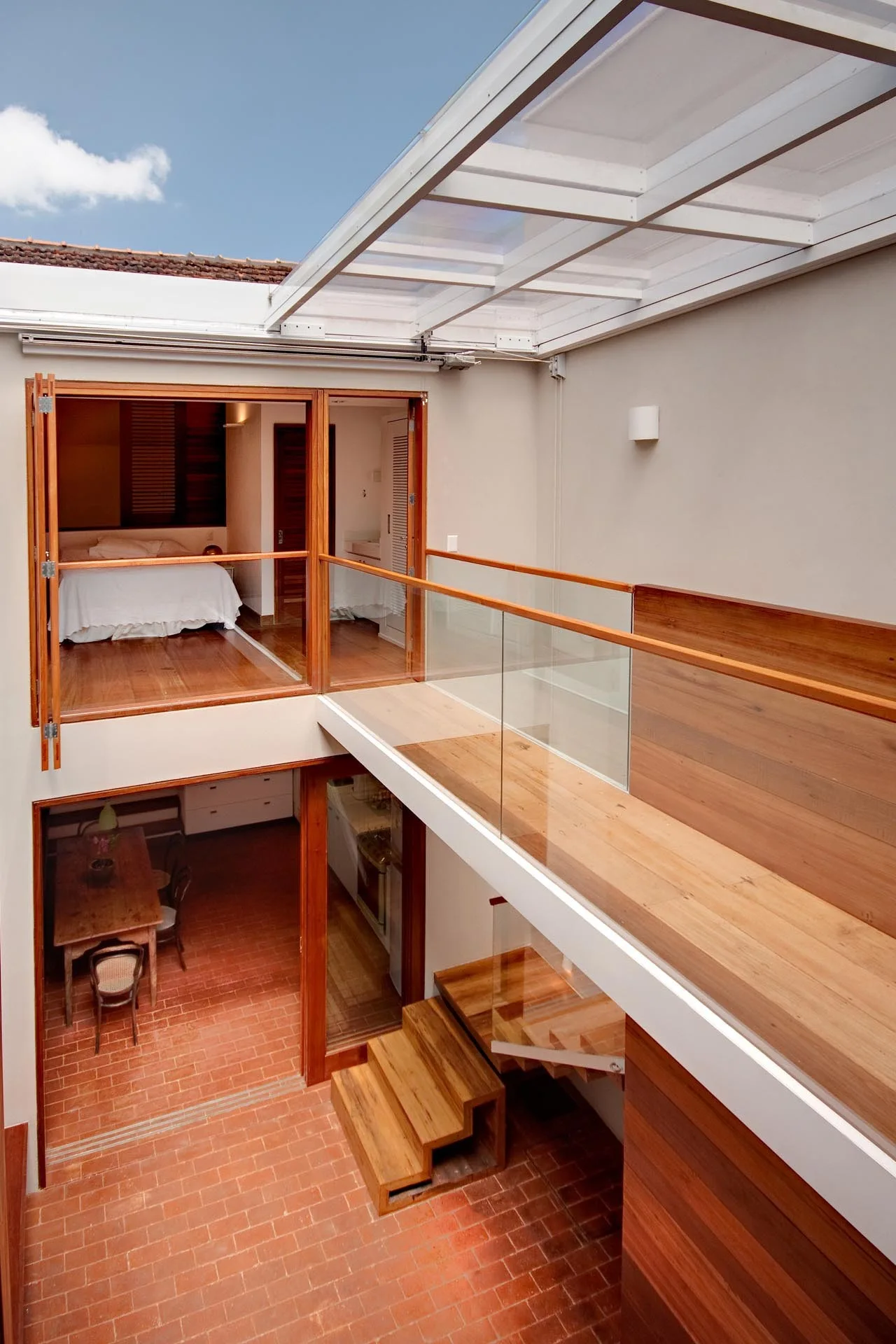 A multi-level interior features a wooden staircase with glass railings under a retractable skylight, overlooking a terracotta tiled dining area.