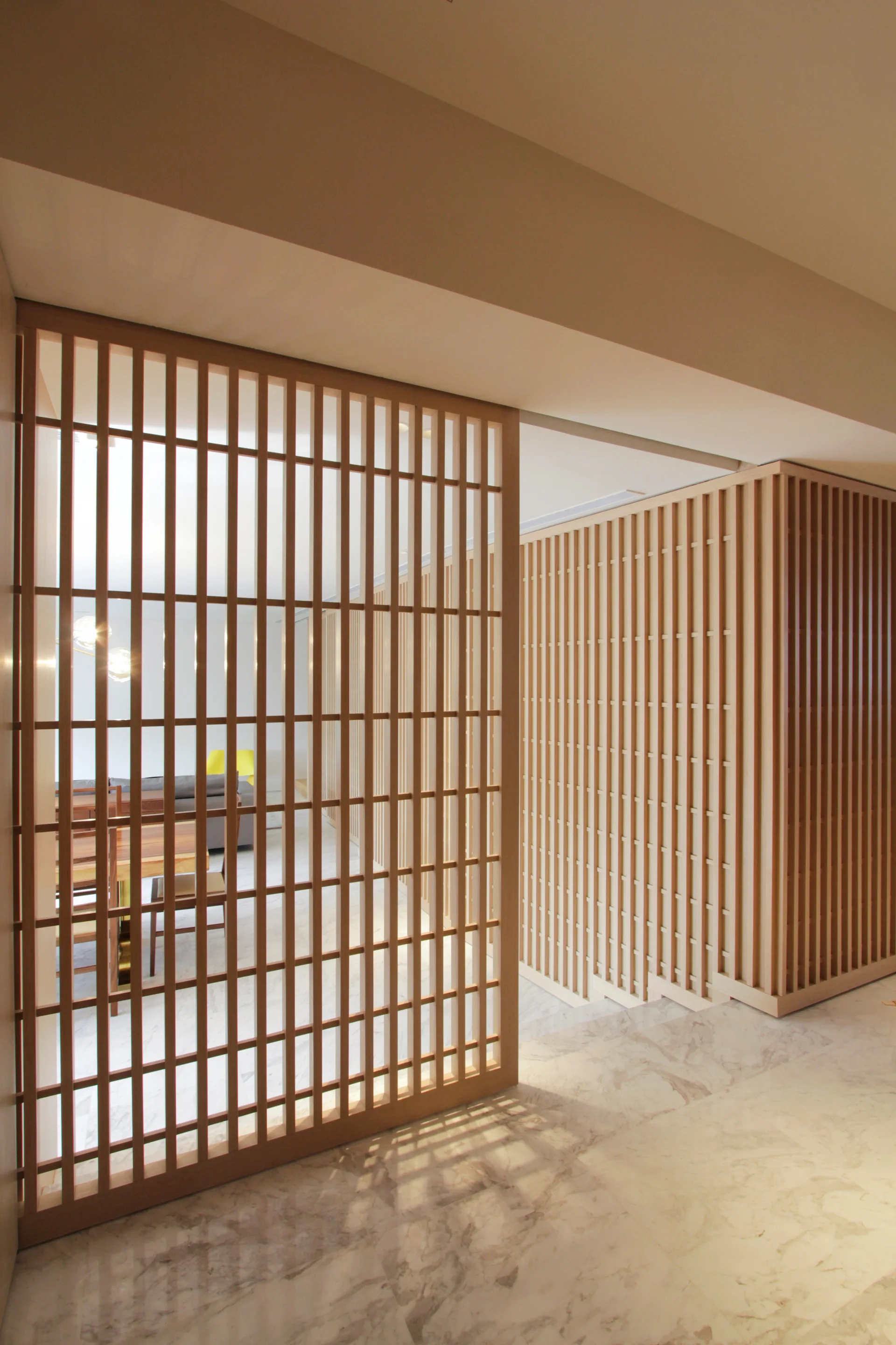 A light wood slatted partition screen stands on a polished white marble floor, revealing a dining table and grey sofa in the background.