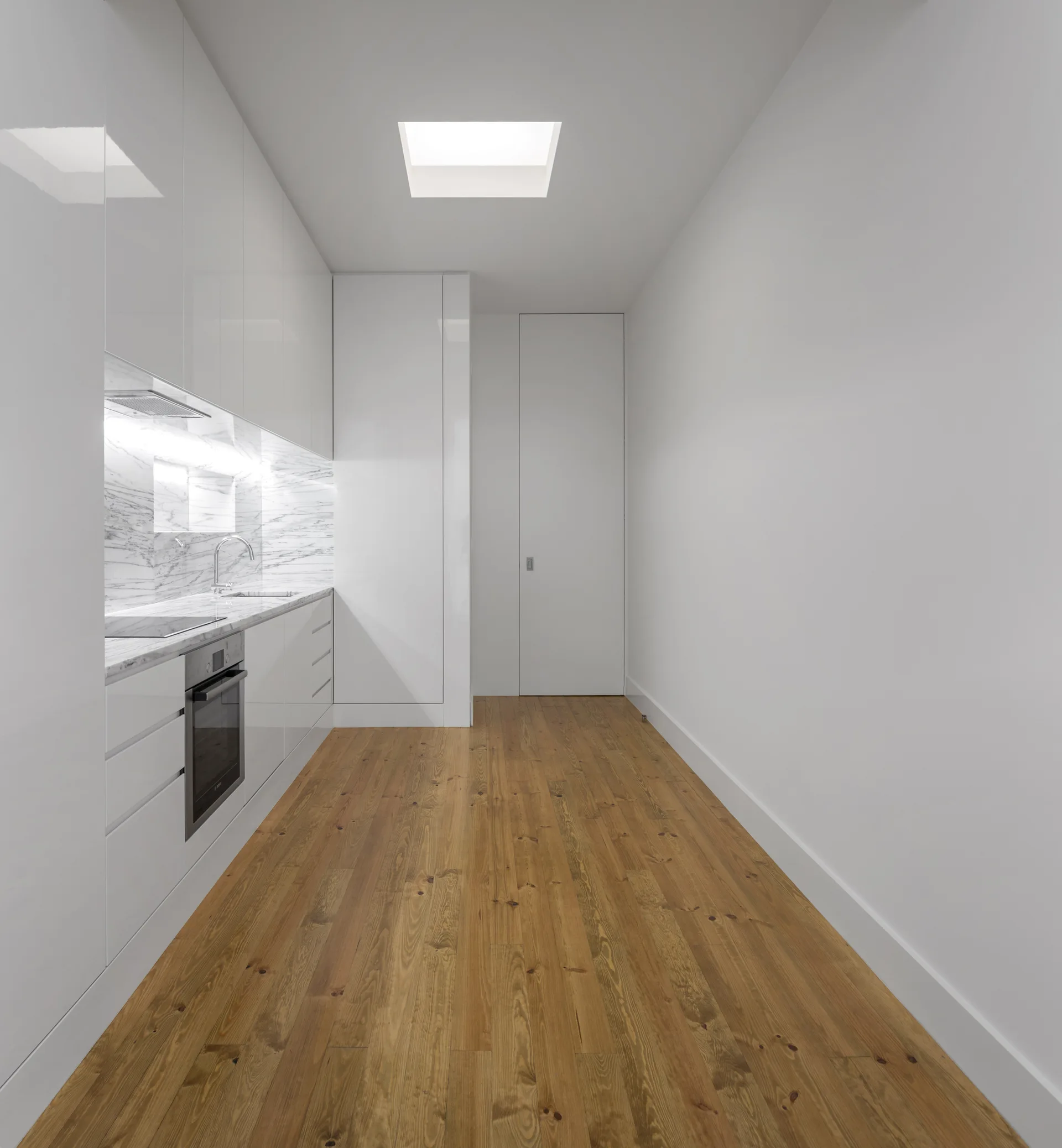 A minimalist galley kitchen features handleless glossy white cabinetry, a marble backsplash, a square skylight, and warm wood flooring.
