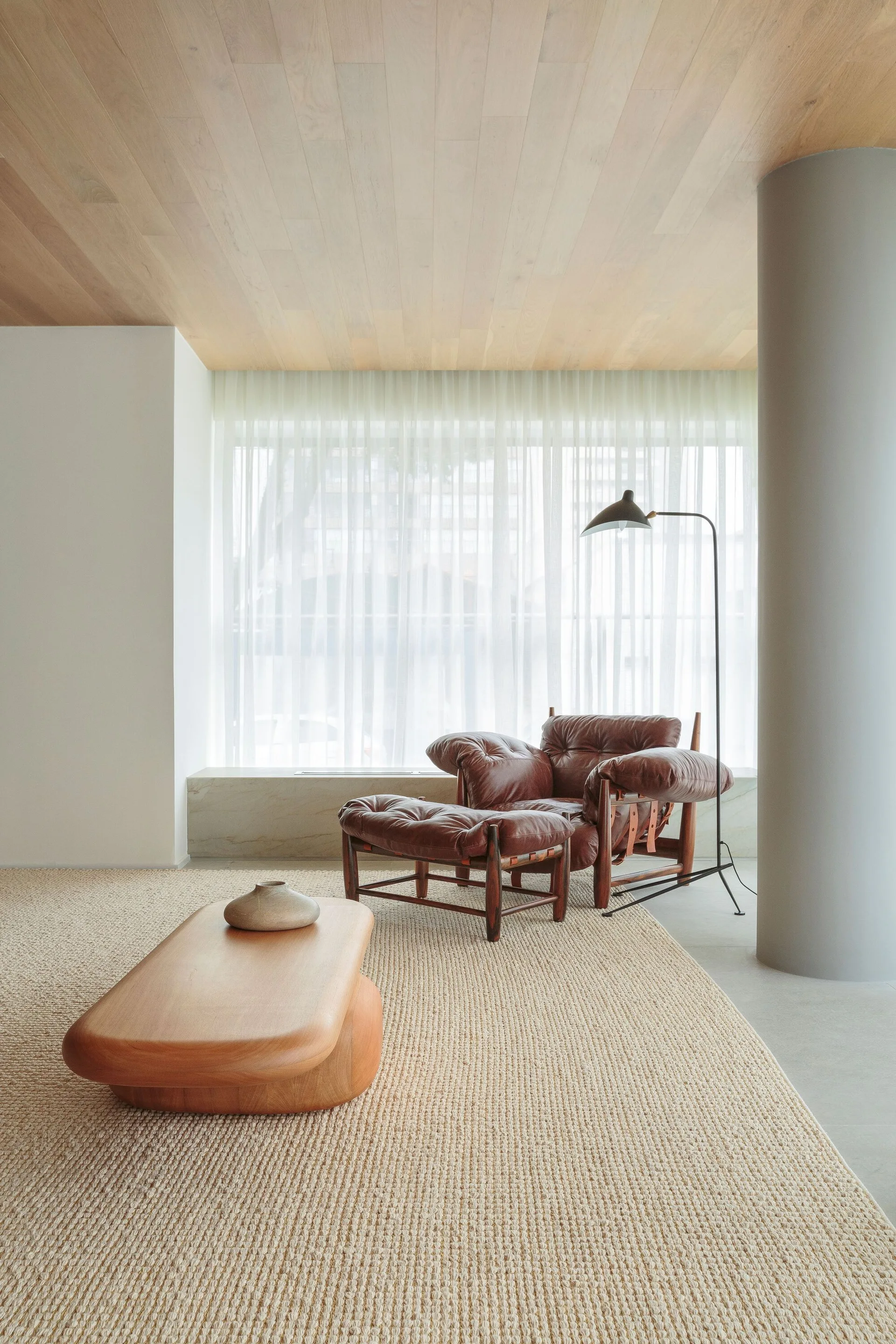 A minimalist sitting area showcases a brown leather lounge chair and ottoman on a woven rug beneath a light wood plank ceiling.