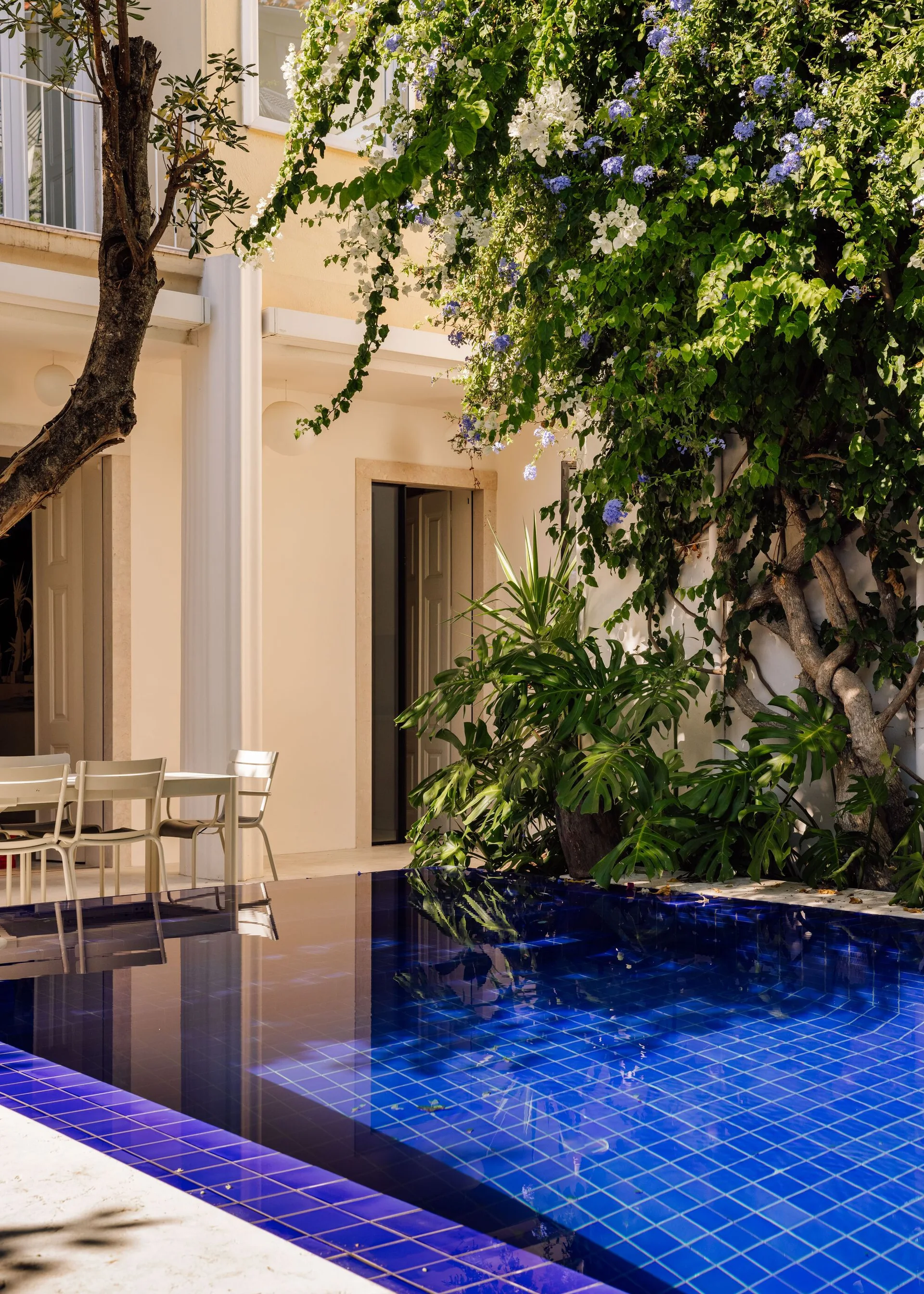 A deep blue tiled swimming pool sits in a sunlit courtyard next to an outdoor dining set and lush climbing vines on beige walls.
