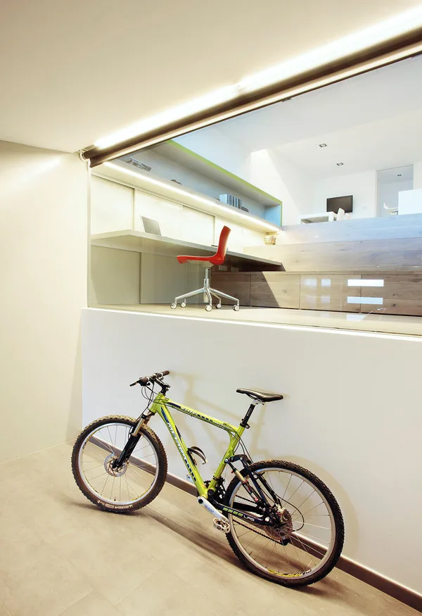 A green mountain bike leans against a white wall below a split-level home office with a floating white desk, red office chair, and glass railing.