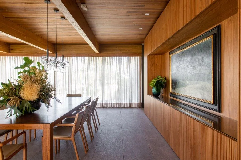 A dining room features a long wood table, matching chairs, floor-to-ceiling sheer curtains, and a wood-paneled wall with a recessed framed painting.