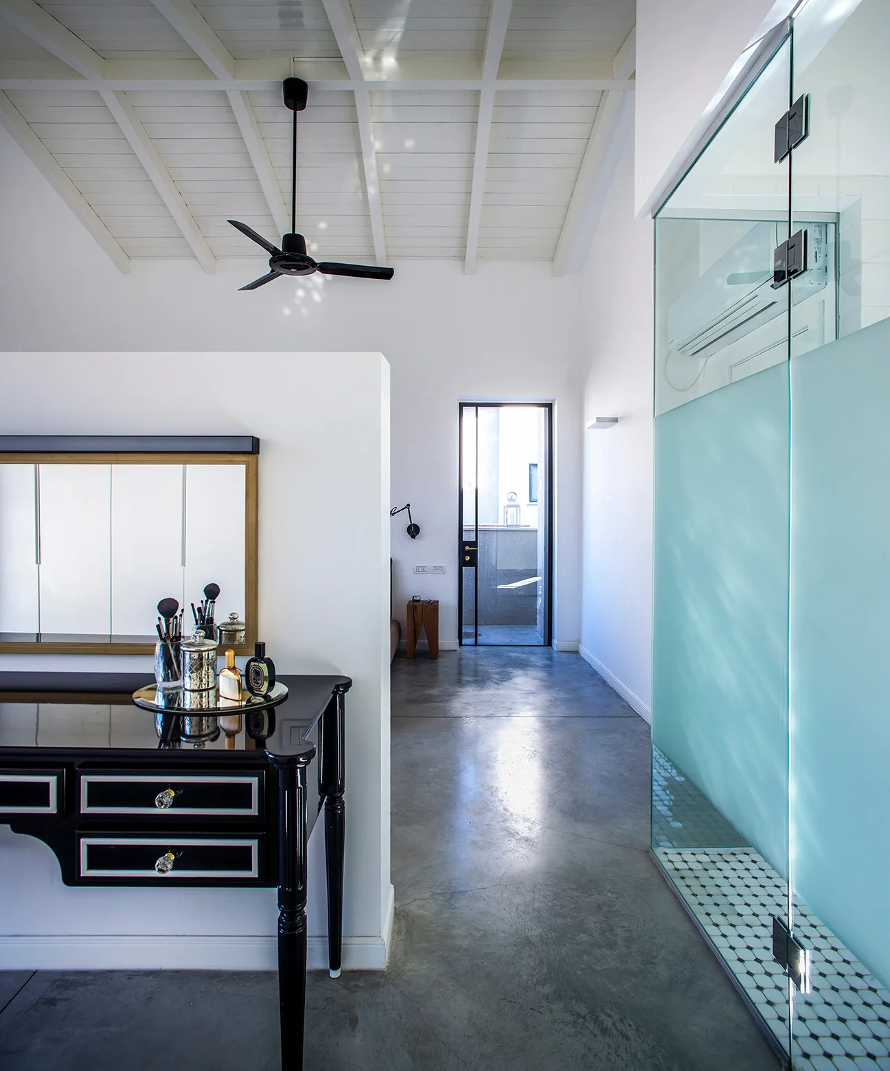 A modern bedroom features a black lacquered vanity desk, a frosted glass bathroom partition, and polished grey concrete flooring under a vaulted white ceiling.
