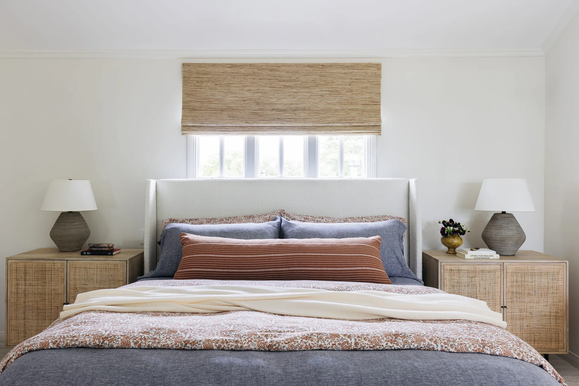 A symmetrical bedroom features a white upholstered wingback headboard beneath a woven shade, flanked by matching cane nightstands with grey ceramic lamps.
