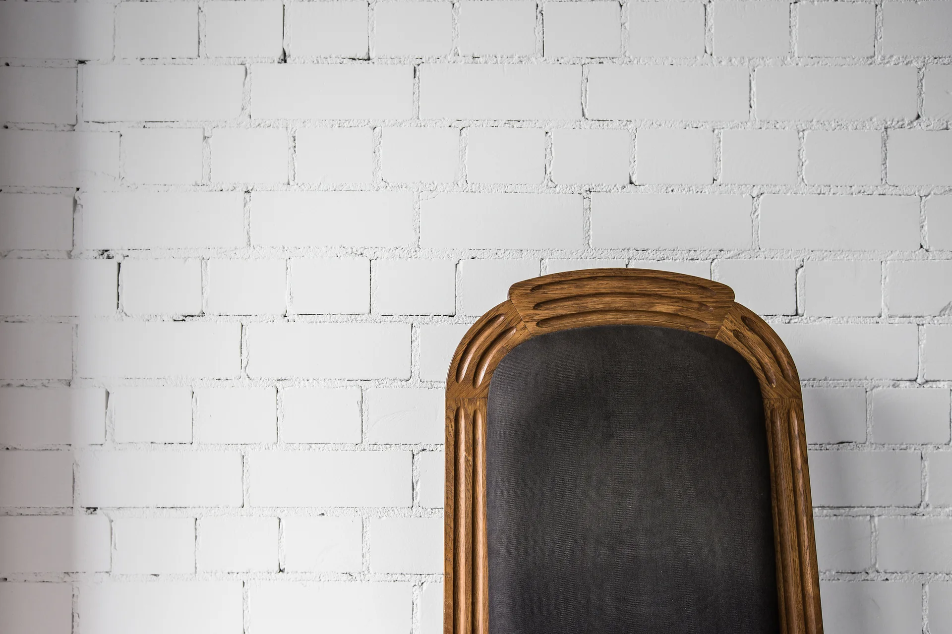 An arched carved wood chair back with dark fabric upholstery sits against a white painted brick wall.