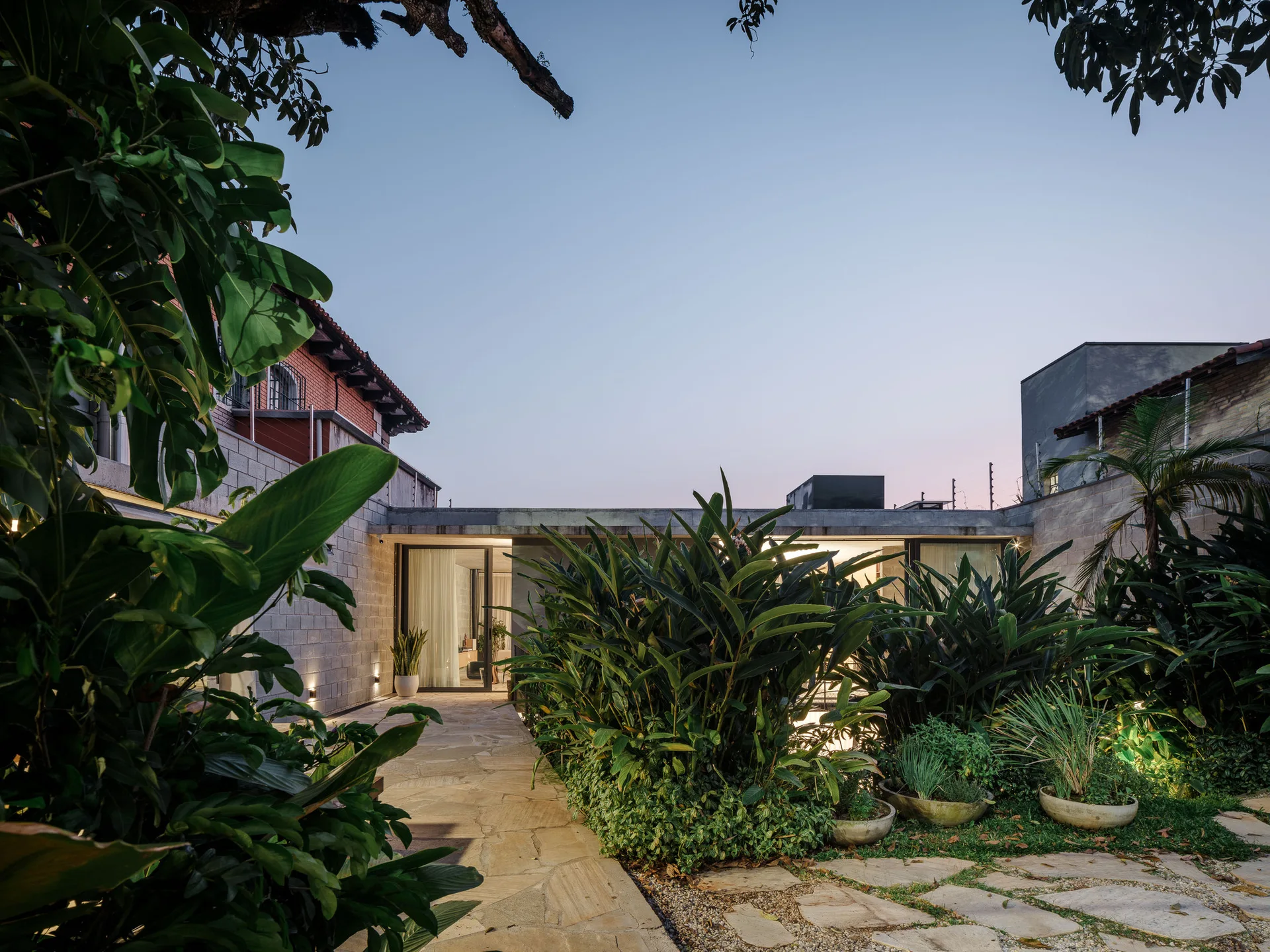 A flagstone path winds past tall green garden plantings toward a modern concrete block house with glowing glass doors at dusk.