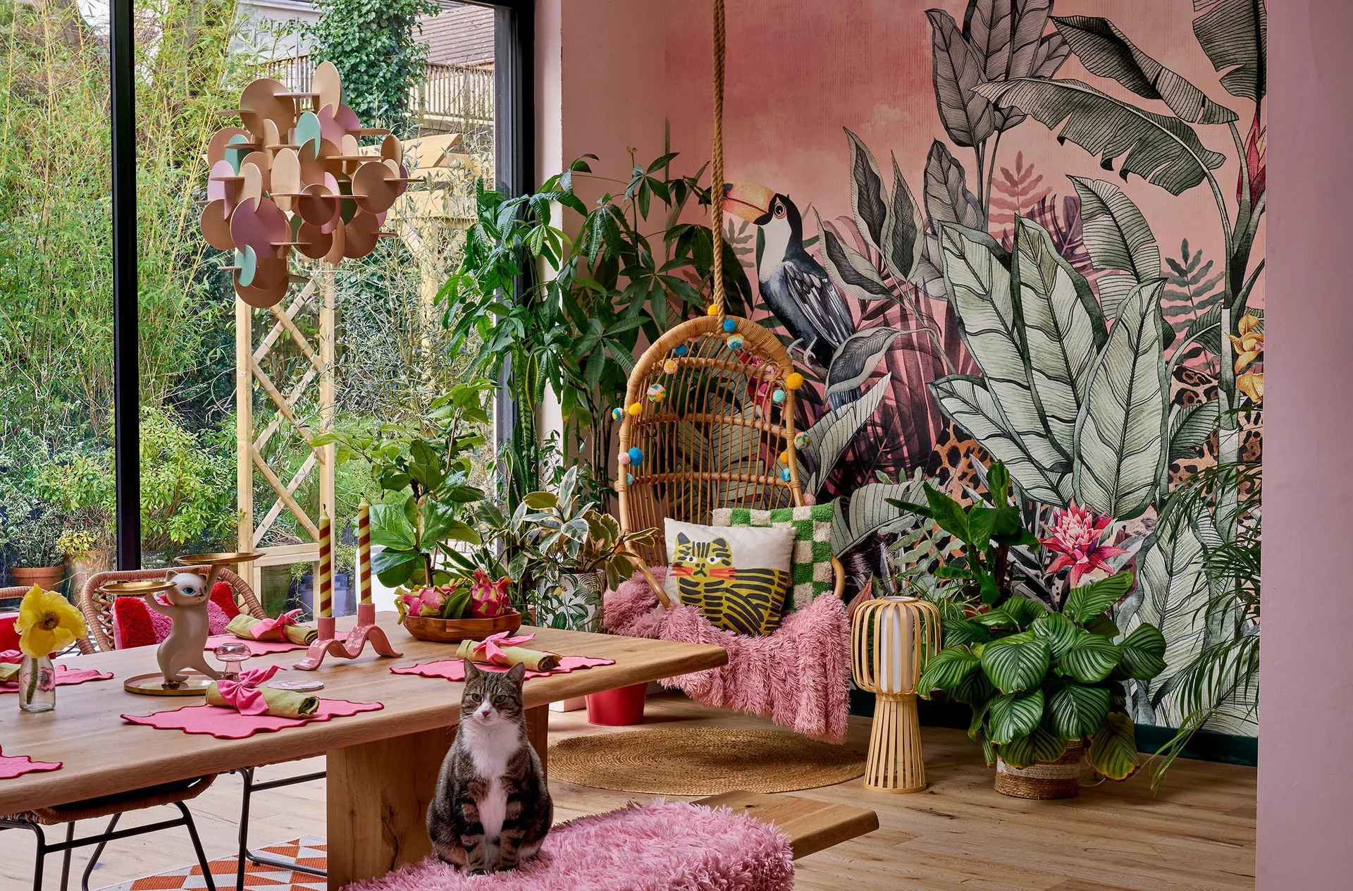 A dining room features a wooden table with pink place settings, a hanging rattan swing chair, and a pink tropical plant mural on the wall.