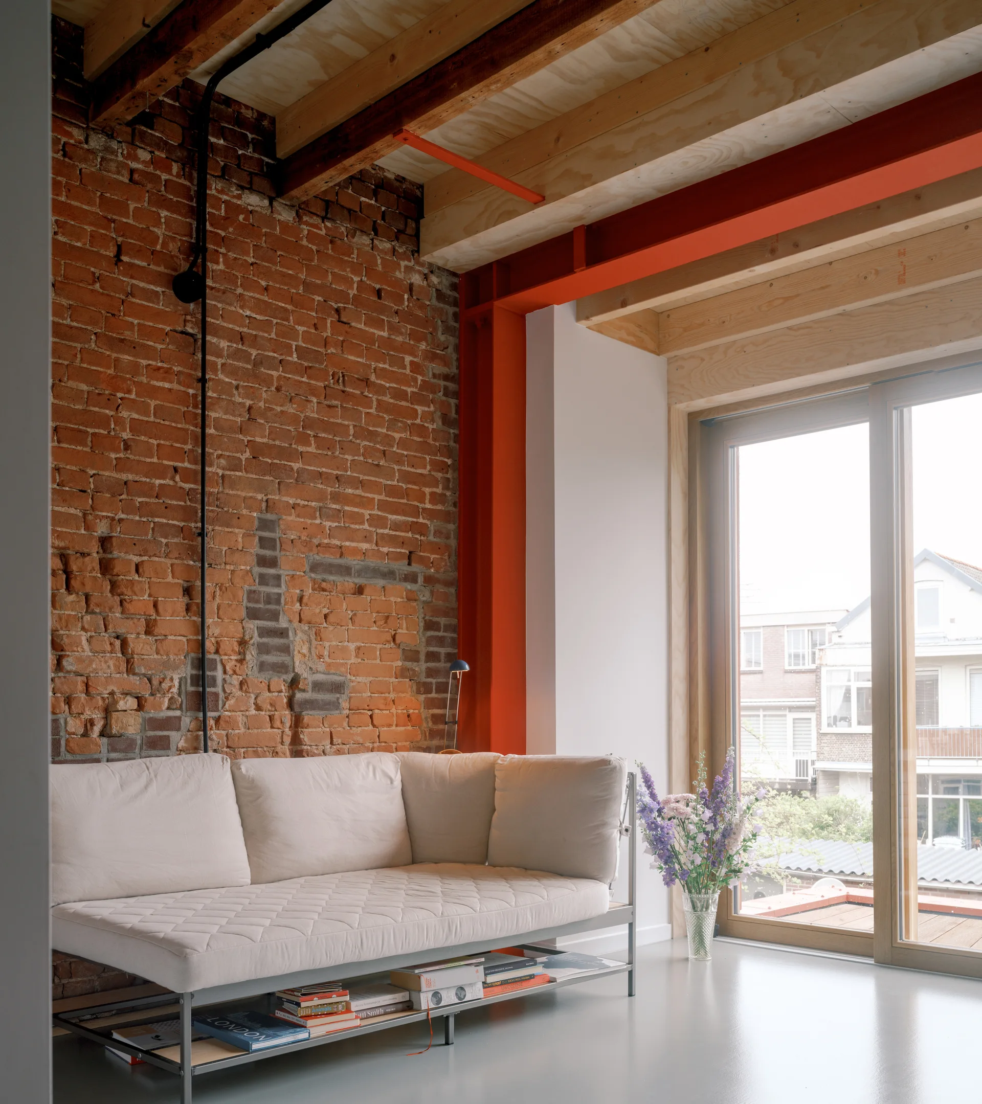 A low cream sofa sits against an exposed red brick wall beneath natural wood ceiling joists and next to a bright orange steel beam.