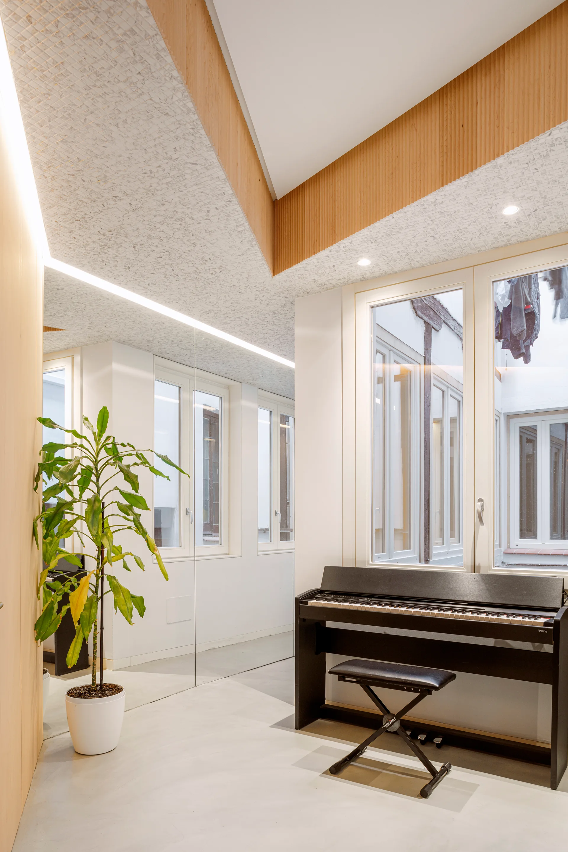 A minimalist music room features a black digital piano, a tall potted plant, and a large mirrored wall below a modern acoustic ceiling.