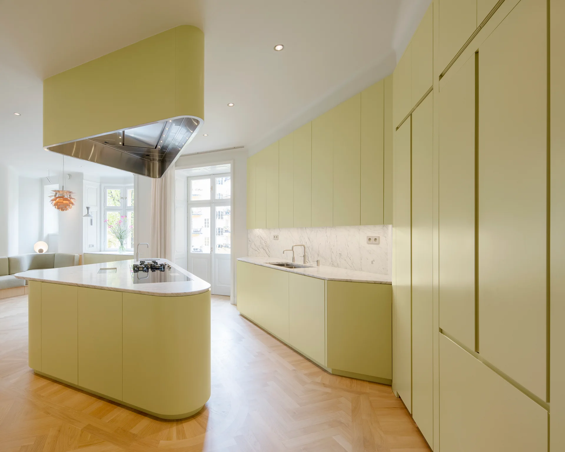 A minimalist kitchen features matte light green cabinets, a curved prep island with a marble countertop, and herringbone wood floors.