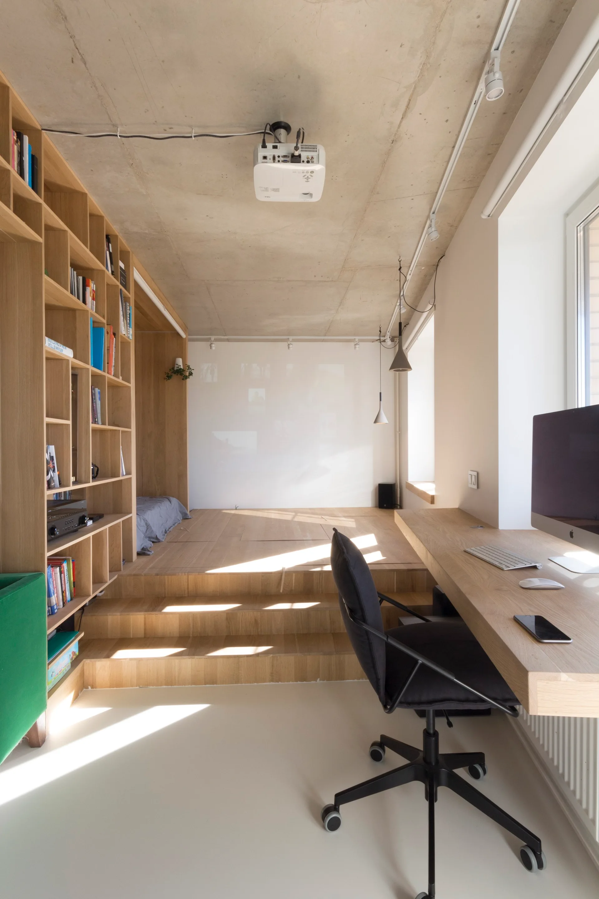 A home office with a floating wooden desk, black office chair, custom wood shelving, and a raised platform floor under an exposed concrete ceiling.
