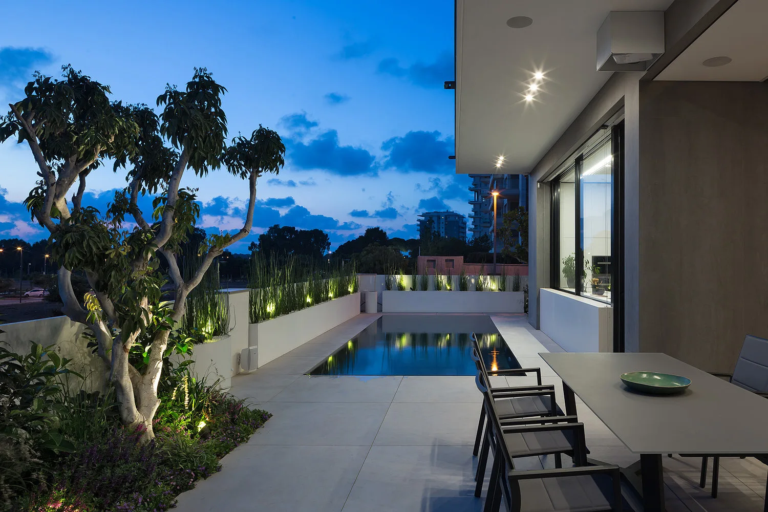 A modern outdoor patio at dusk features a swimming pool, a grey dining table, and built-in white planters with illuminated tall grass.