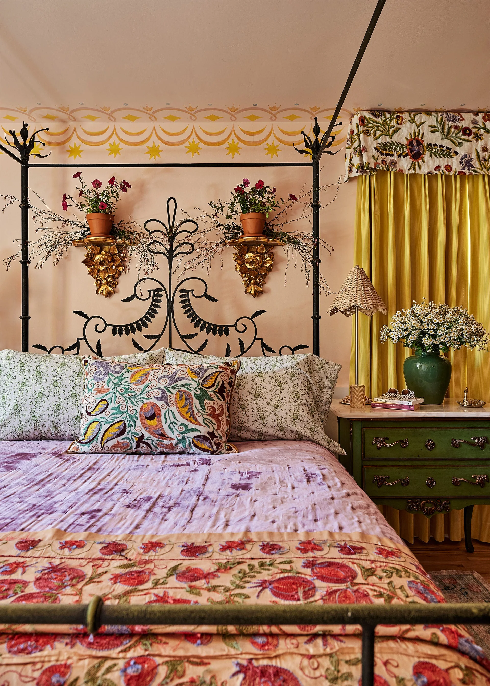 An eclectic bedroom features a black wrought iron bed against pink walls painted with star motifs, next to yellow curtains and a green nightstand.